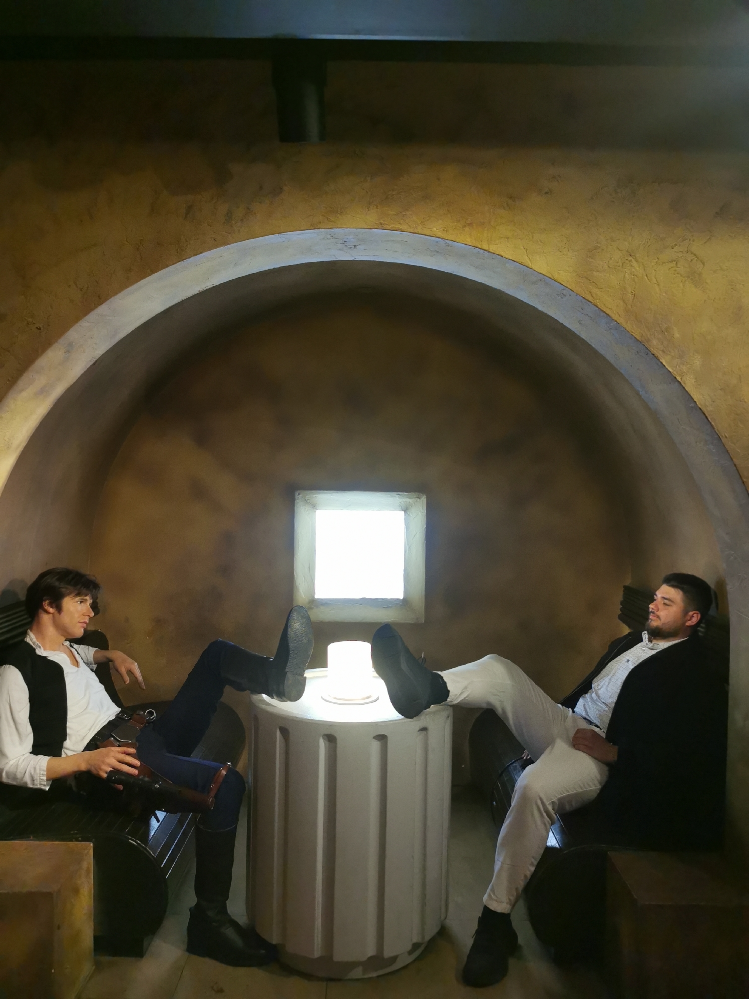 Two men sit facing each other within a room constructed with an arched ceiling and ochre-textured walls. A square window is centered in the back wall, providing bright light. Between the men is a white cylindrical table-like structure with a light source on top.
The man on the left is dressed in attire reminiscent of the character Han Solo from the "Star Wars" franchise, including a white shirt, black vest, dark trousers, and a holster with a prop weapon. He rests his feet on the table, facing the viewer.
The man on the right is dressed in white trousers, a patterned shirt, and a black jacket. He also rests his feet on the table, his body angled toward the viewer.
The setting suggests an indoor recreational or entertainment space, perhaps themed to resemble a location from the Star Wars universe. The scene is not necessarily located in London, United Kingdom.
