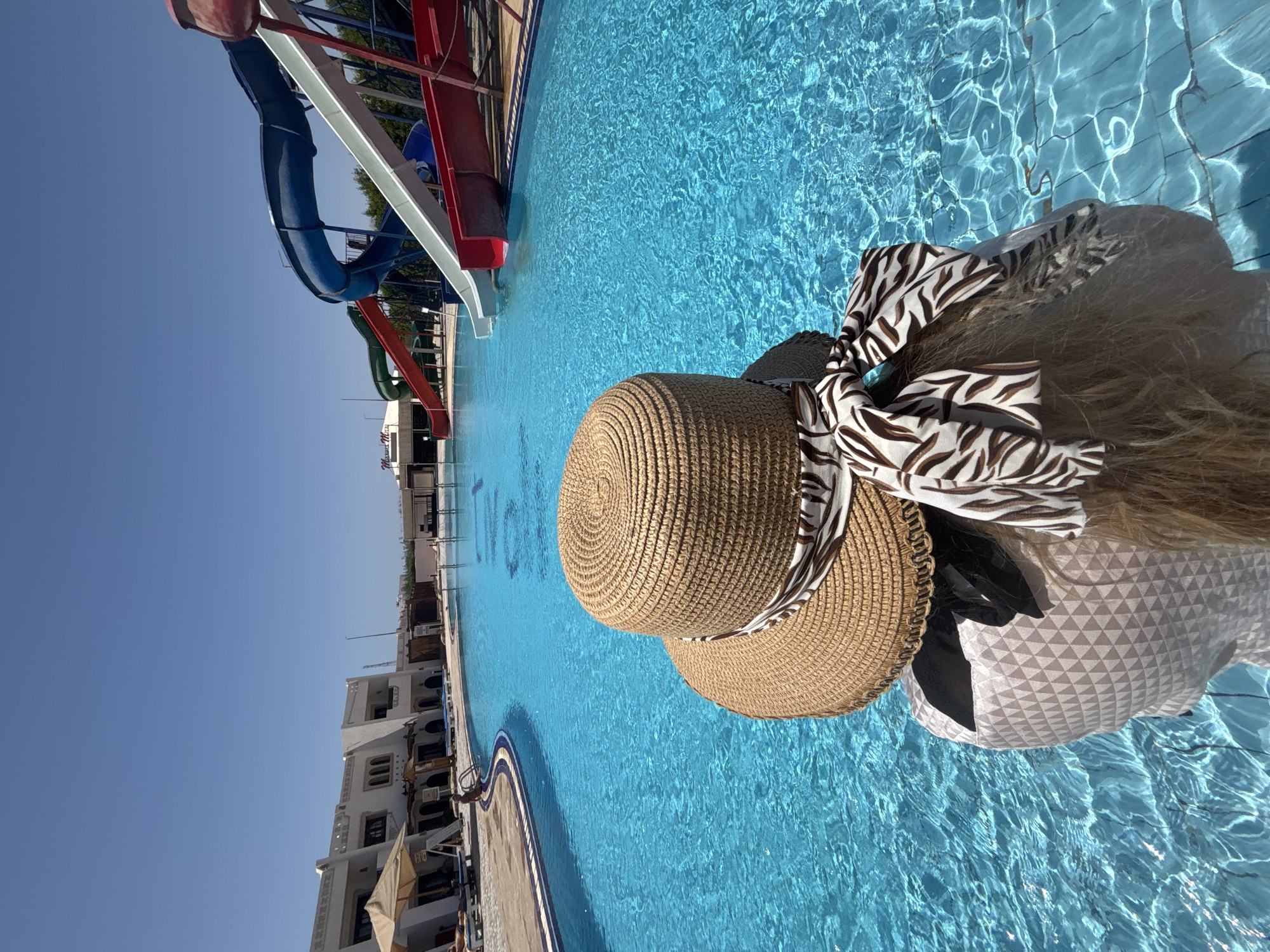 A woman wearing a hat with a zebra-striped ribbon is sitting at the edge of a swimming pool with water slides in the background. The pool is located at a resort with white buildings. The water is clear and blue. The city and country are unknown.