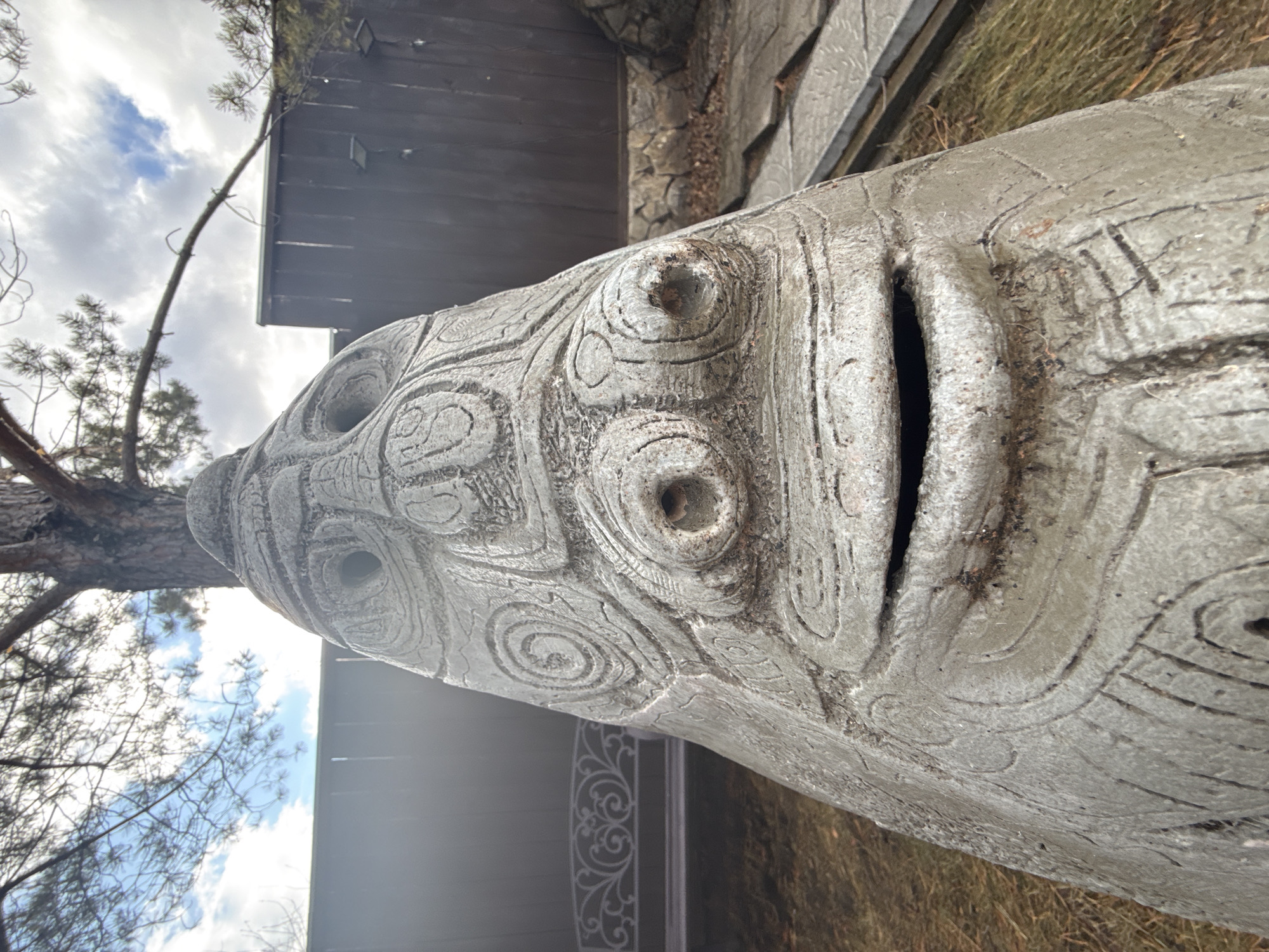 A large, grey, carved wooden statue of a face is situated outdoors, likely in a garden or park. The statue's face features circular carvings for eyes and a deep, dark opening for the mouth. A small tree is partially visible on the left side of the statue, while a dark wooden structure is in the background, possibly a building or fence. The ground is covered with dry grass and other vegetation. The sky above is partly cloudy. No people are visible in the image. There is no information within the image to confirm that the image was taken in Sukhobuzimskoye, Russia.