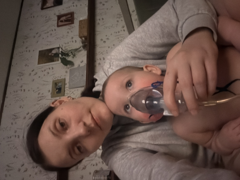 A person holds a baby on their lap, both facing the camera. The baby has a nebulizer mask covering its mouth and nose. The person wears a gray sweatshirt, while the baby has bare legs. The background consists of a wall with multiple framed pictures hanging on it. The scene is casually lit, giving it a personal snapshot quality.