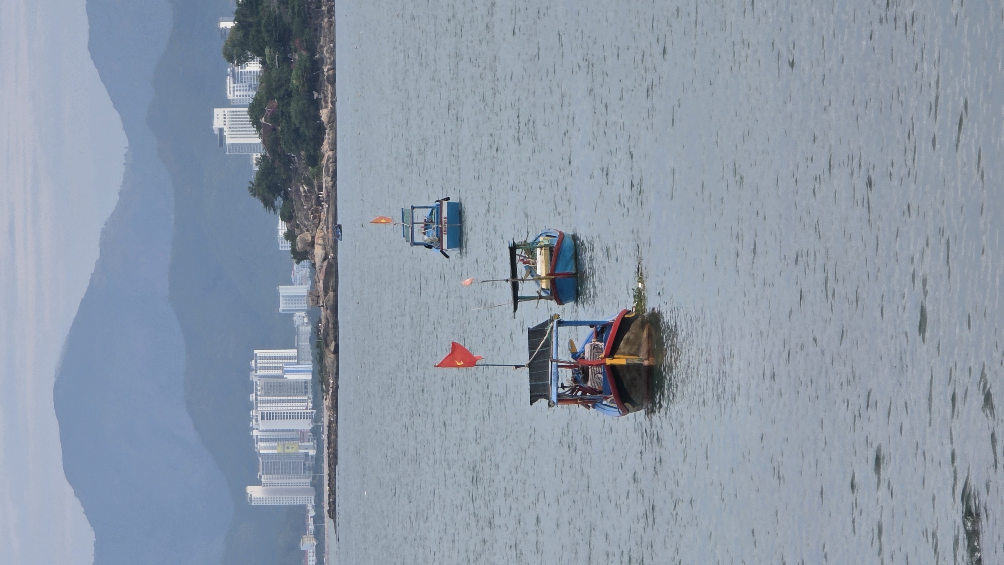 This image captures a serene coastal landscape in Bac Nha Trang Ward, Vietnam, centered on three traditional Vietnamese fishing boats floating on calm, light blue-gray water.
The boats are distinct in color and position. The nearest boat, on the right side of the frame, is a vibrant mix of blue, red, and brown, flying a red flag with a yellow star, a national symbol of Vietnam. Further back, another boat, predominantly blue and brown, also displays a similar red and yellow star flag. The third boat, furthest in the background, is primarily blue, with the name "ANH VAN" clearly visible in white letters on its side. These are small, wooden vessels, likely used for local fishing or transport, equipped with simple structures and poles.
The background features a picturesque coastline with a mix of natural and urban elements. A rocky shore, dotted with green trees and foliage, stretches along the water's edge. Behind this, several modern, multi-story buildings with white exteriors are visible, interspersed among the greenery, indicating a developed area. Further in the distance, a range of large, hazy blue-gray mountains rises, creating a soft, layered backdrop.
The scene appears to be set during the daytime, under soft, diffused lighting, possibly on an overcast day or during a time of day when the sun is not directly overhead. The water shows gentle ripples, suggesting a calm bay or harbor. There are no overt human activities clearly visible, but the presence of the fishing boats suggests maritime life is central to this location.
