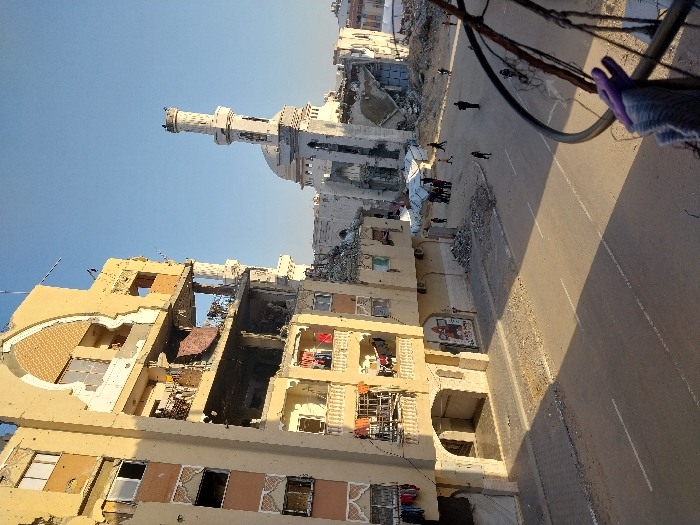 This image depicts a devastated urban streetscape in Khan Yunis, Palestinian Territory, under clear blue skies. On the left, a multi-story beige building, approximately seven floors high, exhibits extensive structural damage. Multiple floors have sections of their exterior walls collapsed or blown out, exposing interior rooms. Balconies on the lower-middle floors show hanging laundry, including red, black, and white garments. The building's facade features decorative archways and patterns, with visible signs of impact damage or decay.
In the mid-ground, a white mosque is severely damaged. Its prominent minaret, with a distinct balcony near the top, remains largely standing but shows wear. The main body of the mosque's structure, including its dome, is partially collapsed, with significant rubble surrounding its base. Adjacent to the mosque, several white tents are erected amidst debris.
The foreground features a paved asphalt street with white lane markings, flanked by concrete sidewalks. Long shadows are cast across the road. Scattered debris and rubble are visible on the sidewalks, particularly near the damaged structures. Approximately 10-15 individuals are present on the street and near the mosque's vicinity; some are walking, and a small group is gathered near the white tents. The perspective suggests the photograph was taken from an elevated position, with a blurred metal railing and a piece of purple fabric visible in the bottom right corner.
