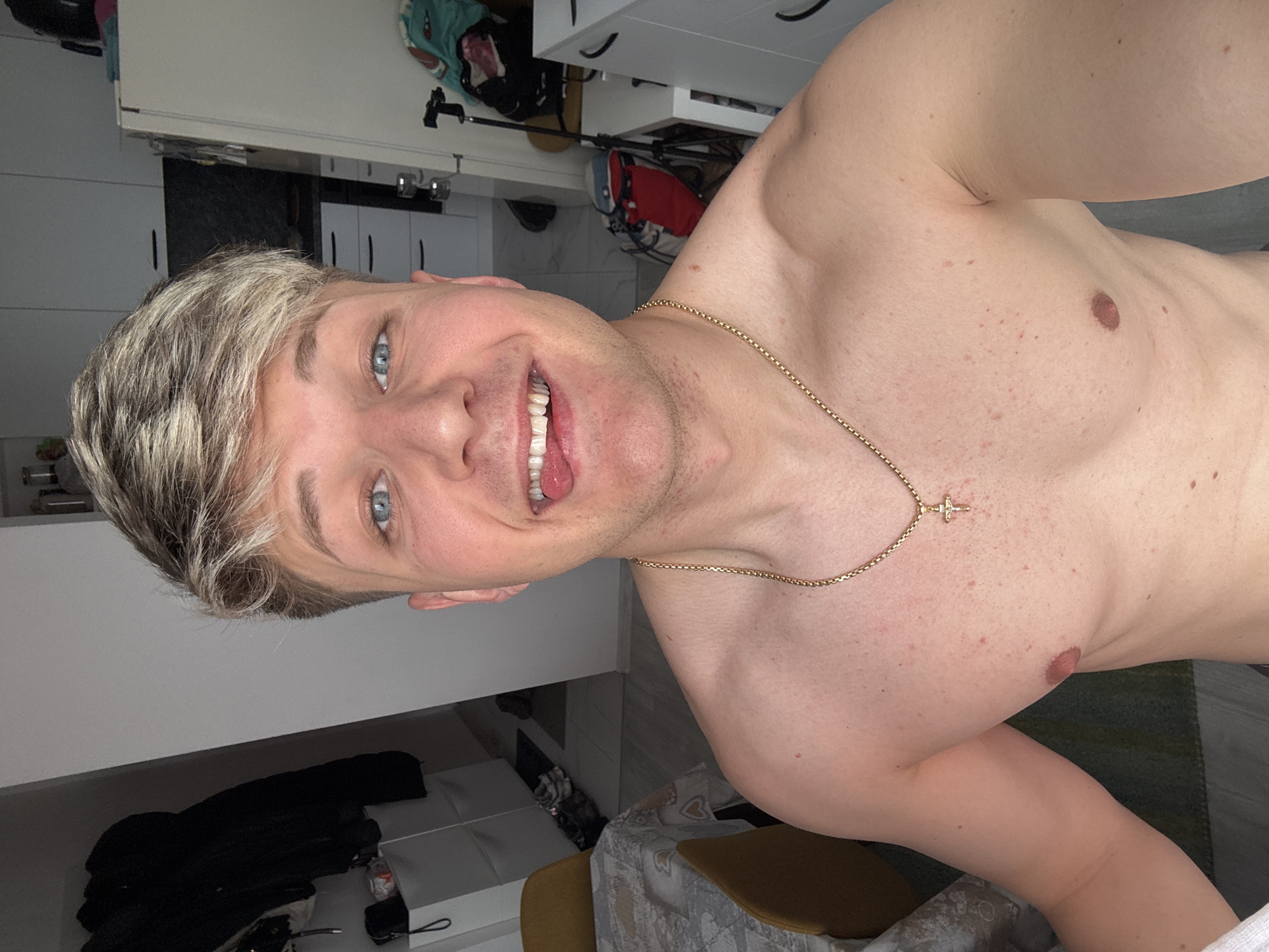 A light-skinned male with bleached blonde hair is smiling and wearing a gold chain with a cross pendant. He is shirtless. The background includes a white kitchen, a chair with a patterned tablecloth, and personal belongings. The location cannot be determined as Plovdiv, Bulgaria, based on the provided image.