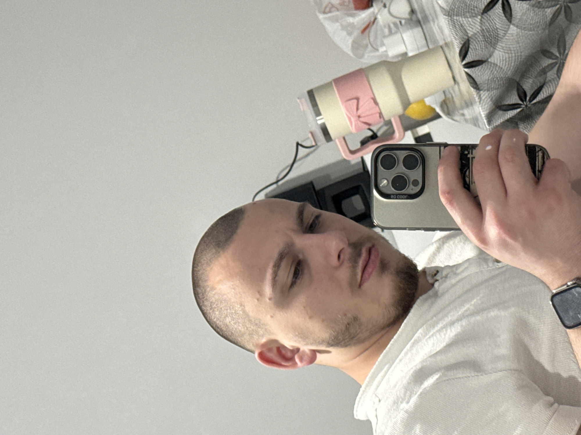 A man with a closely shaved head and a beard is holding a smartphone in front of him, presumably taking a selfie. He is wearing a white collared shirt and a watch. The background includes a white wall, a gray and white patterned fabric, a beige and pink thermos, and some other indistinct objects. The image does not offer any information to determine if the man is located in the city of Stara Zagora, Bulgaria.