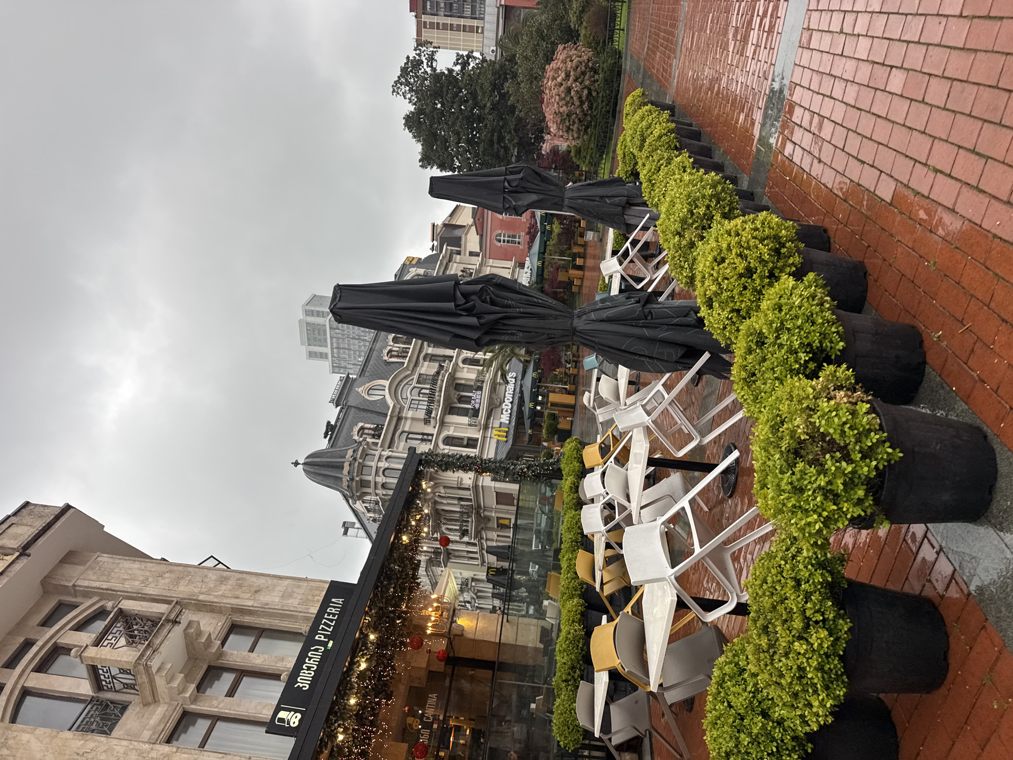 This media file captures an outdoor urban scene, likely a street or plaza, on a visibly overcast and damp day. The ground, especially the brick-paved path on the right, appears wet, suggesting recent rain. No people are present, lending a quiet and somewhat deserted atmosphere to the setting.
In the foreground and midground, the outdoor seating area of a cafe or pizzeria is visible. Numerous chairs, predominantly white with some yellow accents, are stacked or arranged, indicating the area is not currently in use. Several large, black, folded outdoor umbrellas stand ready for deployment. These elements are arranged alongside a row of neatly trimmed green bushes planted in black pots, separating the seating area from a reddish-brown brick pathway that recedes into the background on the right.
On the left, the entrance to the cafe is adorned with an awning featuring string lights and a few red decorative baubles. A sign on the building clearly displays "ЗАВОД PİZZERİA", with Cyrillic script (ЗАВОД) followed by "PIZZERIA". Below this, "CANTINA" and "360" are partially visible.
The background is dominated by several multi-story buildings showcasing diverse architectural styles. A notable structure features a distinctive dark roof, a turret-like dome, and ornate detailing. Further down the street, a prominent yellow "McDonald's" sign is visible, along with other commercial signs and trees. The sky is uniformly gray and cloudy, providing diffused, dim lighting to the entire scene.