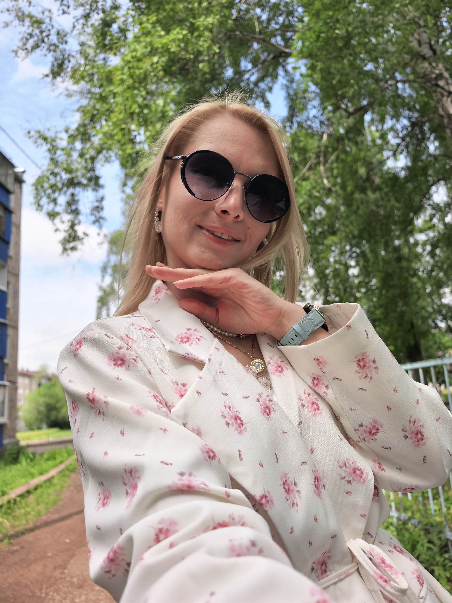 A blonde woman takes a bright, upward-angled selfie outdoors in Sterlitamak, Russia, on a sunny day. She is smiling and looking directly at the camera, with her left hand gently resting on her chin. She wears large, round dark sunglasses, pearl drop earrings, and a layered necklace consisting of a gold circular pendant and a pearl choker. Her clothing is a white long-sleeved top or blazer with a delicate pink floral pattern. A watch with a light blue leather strap is visible on her left wrist. The background features a clear blue sky partly covered by lush green tree foliage, indicating good weather. To her left, a multi-story building with blue details and a reddish-brown path are visible. A light-colored metal fence can be seen on the right side of the image. The scene appears cheerful and relaxed. There is no visible text.