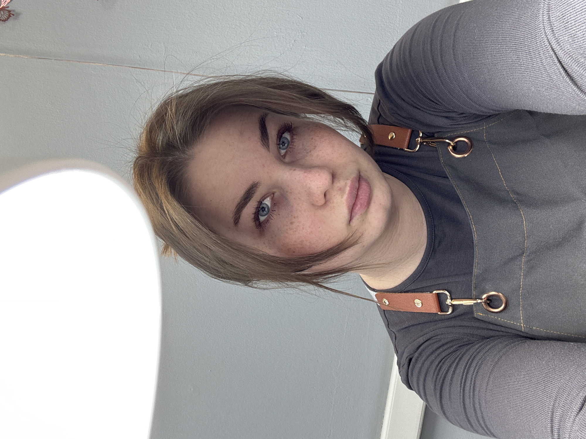 An indoor, close-up, chest-up portrait depicts a young adult female located in Chaikovsky, Russia. The individual has fair skin with visible freckles across her nose and cheeks, blue eyes, and light brown hair with some strands falling forward. She is wearing a dark gray long-sleeved shirt beneath a dark gray apron. The apron features brown faux leather straps with gold-colored metal hardware, including buckles, snaps, and rings, visible on both shoulders. The subject maintains direct eye contact with the camera, displaying a neutral expression. The background consists of a light gray, textured wall. A large, bright, white, semi-circular light fixture is visible in the upper left of the frame. A thin, light-colored string hangs vertically in the upper right, and faint red butterfly-shaped decorations are discernable on the wall in that area. A white horizontal trim is present along the bottom edge of the wall in the left background.