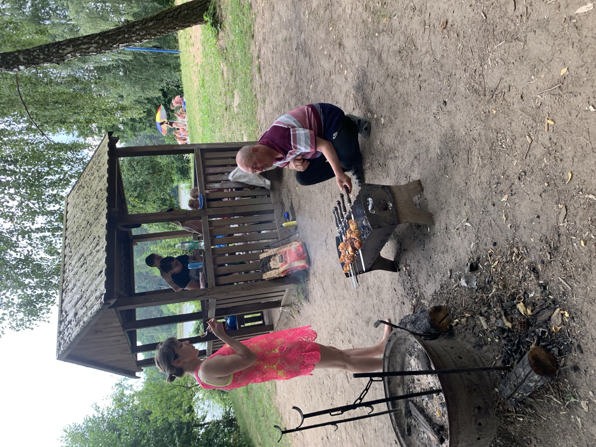 In the village of Grozovo, Belarus, a man is crouched by a grill, tending to skewers of meat. He wears a striped shirt and dark pants. Nearby, a woman in a vibrant pink dress stands, holding a fork, and appears to be eating. Behind them, a wooden gazebo stands, with several people visible inside. The ground is dirt and sand, with scattered debris. To the left, a large metal fire pit is visible, with logs and charcoal around it. In the background, trees and a body of water suggest a natural setting, with other people visible further in the distance enjoying the outdoors.