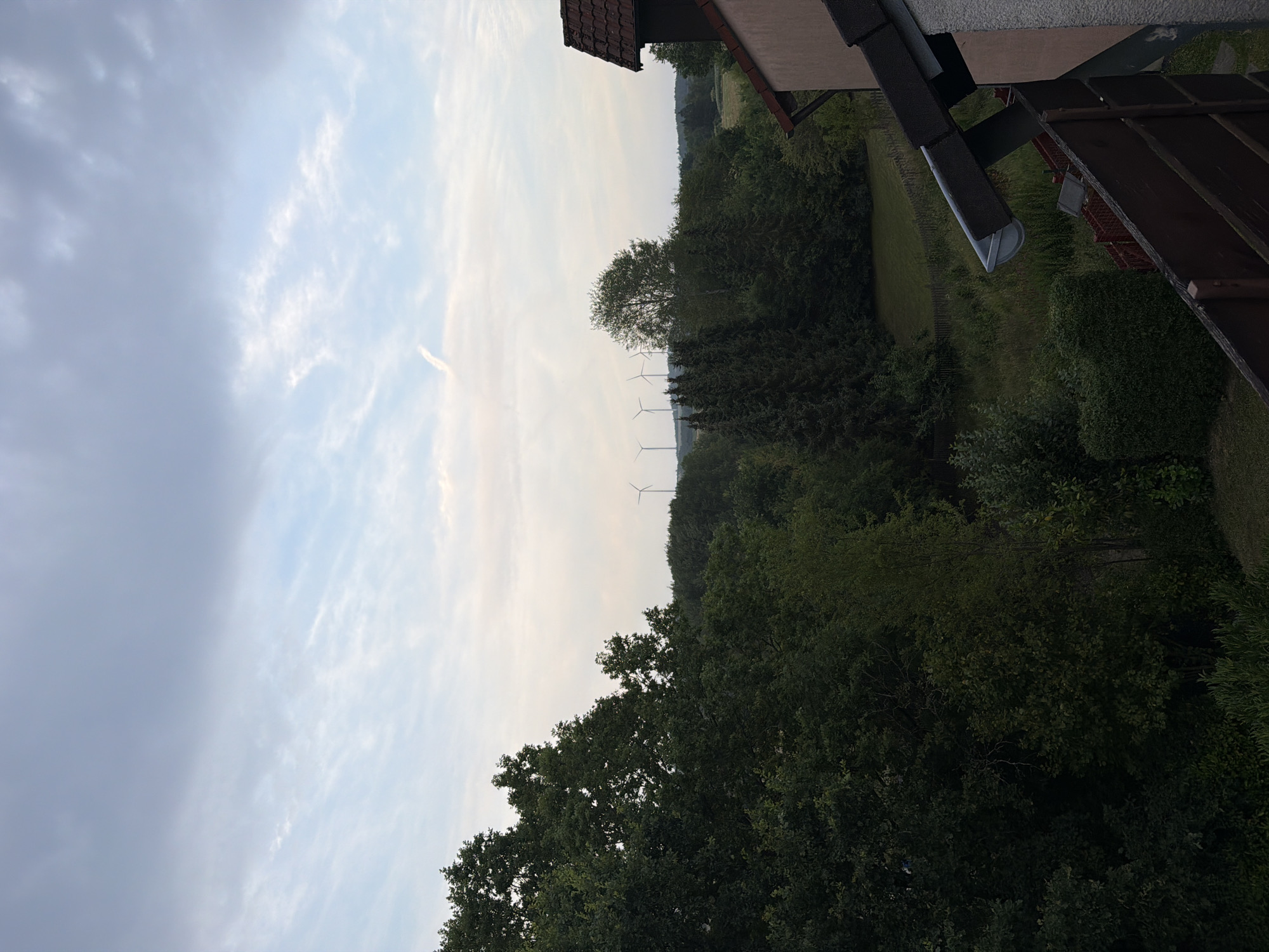 A wide-angle, high-angle shot depicts a scenic view from a balcony in Creußen, Germany. The foreground is dominated by a dark brown wooden railing and a portion of a light-colored building with a terracotta-tiled roof. Beyond the railing, a lush green lawn stretches out, bordered by a wooden fence and dense trees. In the middle ground, a line of various trees, including coniferous and deciduous varieties, creates a natural barrier. In the background, a field with some scattered trees is visible, and on the horizon, several wind turbines stand tall against a cloudy, pale blue sky. A faint contrail is visible in the sky. The overall scene suggests a peaceful, rural or semi-rural setting with a focus on natural elements and renewable energy sources.
