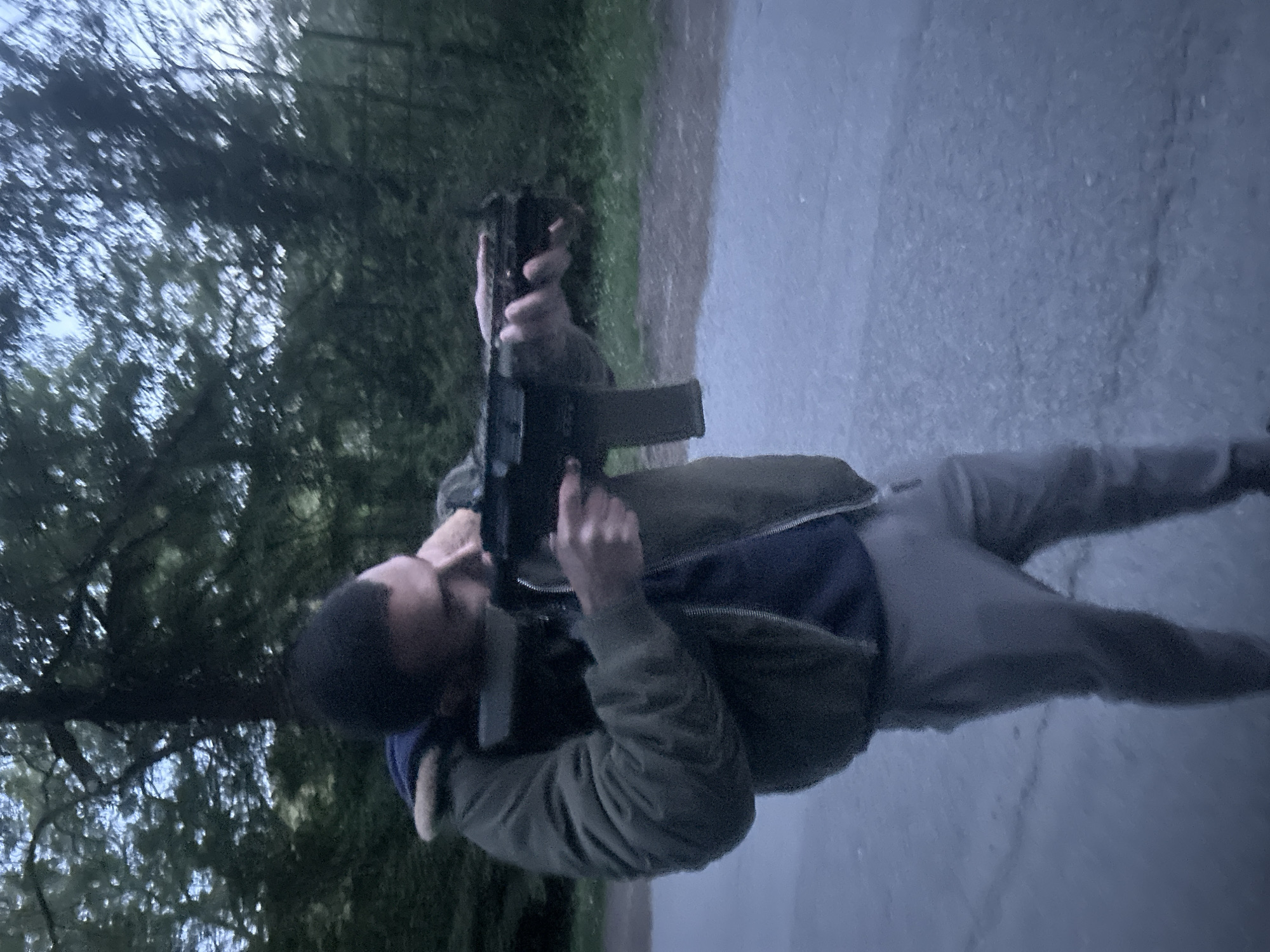 A person is holding a firearm. The person is wearing a dark green jacket over a dark blue top with light grey pants. The person is standing on pavement next to some green vegetation. The photograph appears to be taken outside during the day with low light. It is not possible to determine the location of the scene or the intent of the person with the firearm.