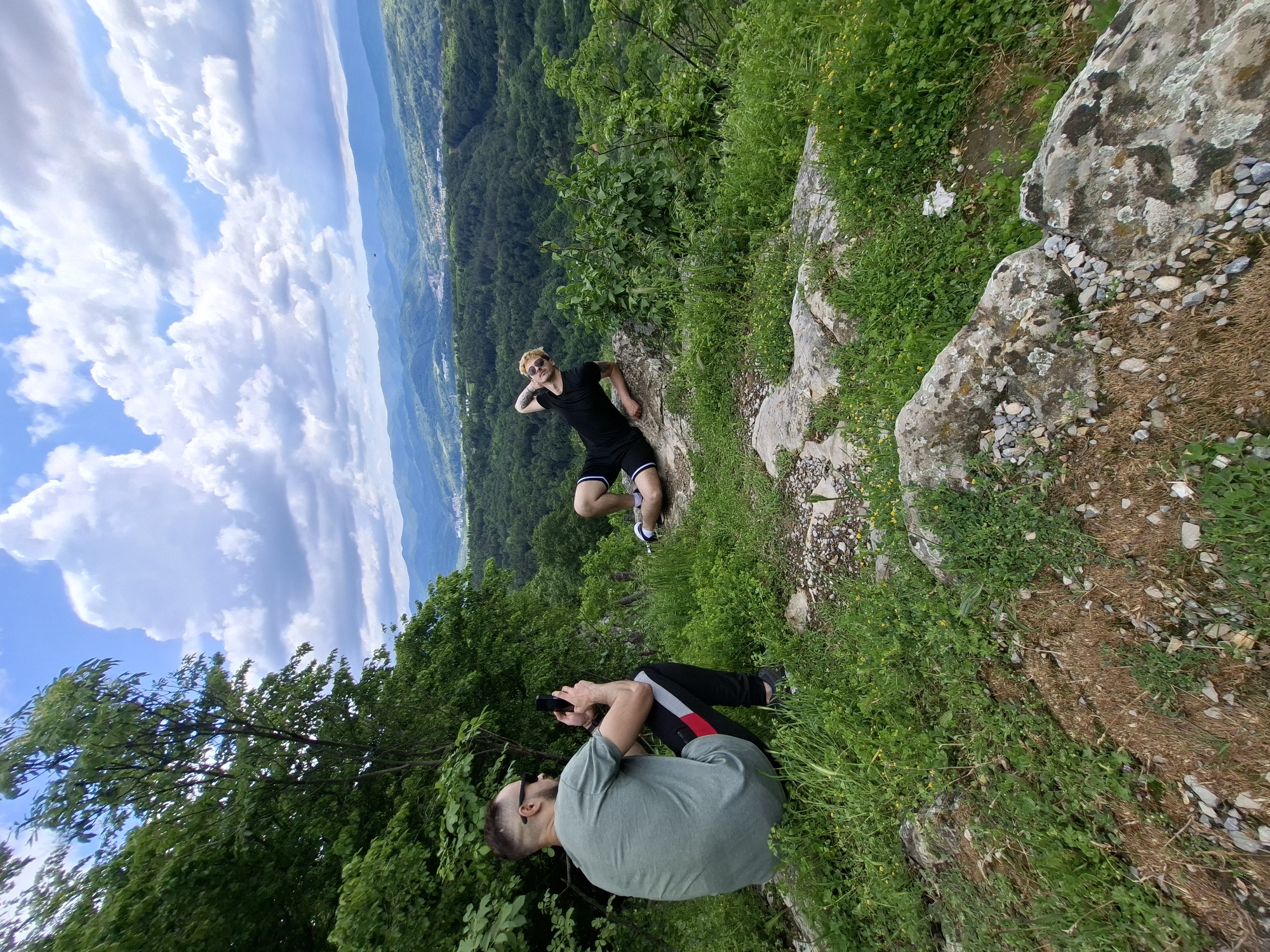Two men are positioned on a rocky, grassy outcrop. One man, wearing a light green t-shirt and dark pants with red and white stripes, is seated and holding a black mobile phone in his hands. He is also wearing sunglasses. The other man, wearing a black shirt and black shorts with white stripes, is leaning back on the rocks with one hand on his head and sunglasses on. The surrounding area is mountainous, with a clear view of Gabrovo, Bulgaria, nestled in the valley below. The sky above is blue with puffy white clouds. Vegetation is abundant, consisting of trees and grass on the hillside. Rocks and small stones are scattered around the grassy area.