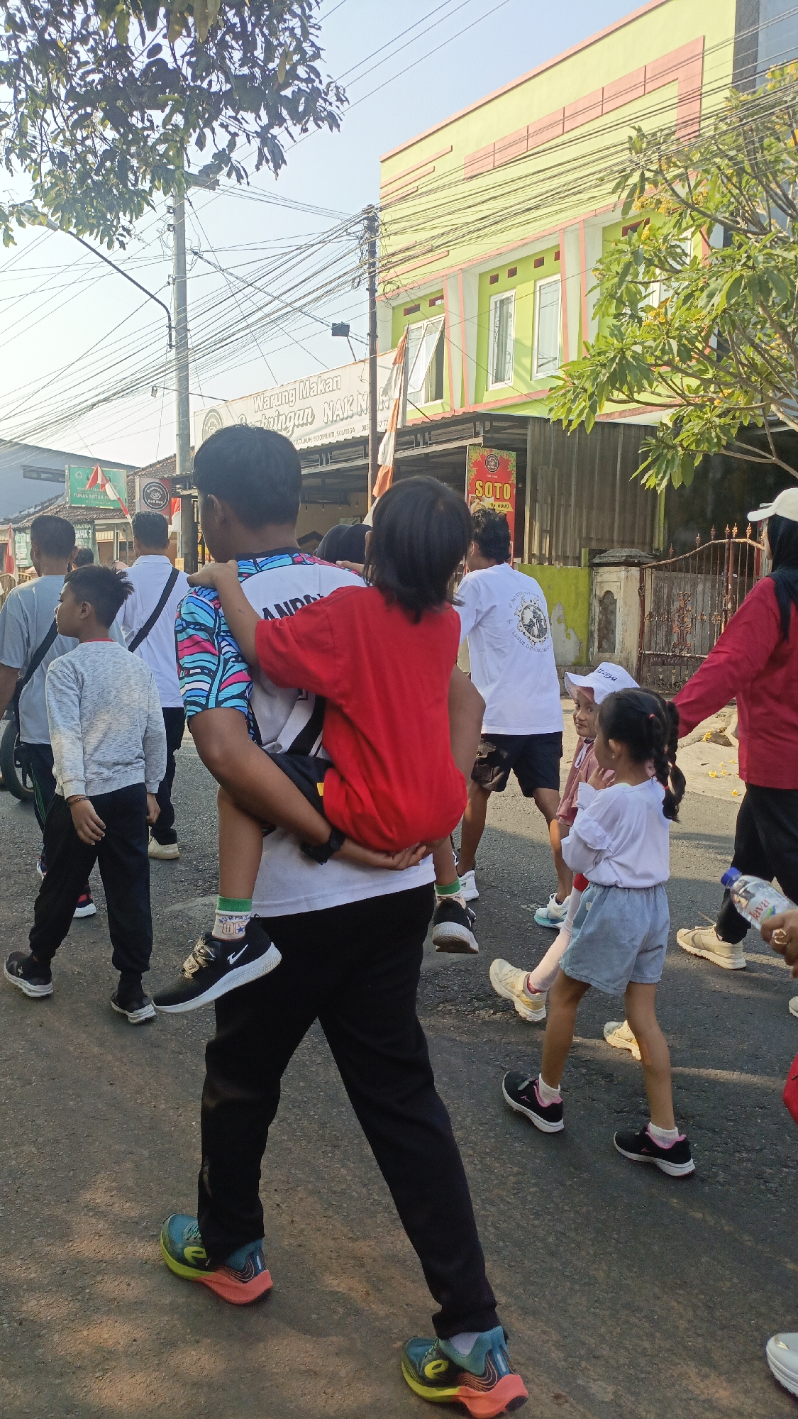 The image depicts a street scene in Salatiga, Indonesia, likely during a public event or procession. In the foreground, a young boy is being carried on the shoulders of an older boy. The older boy wears a white shirt with a colorful, abstract pattern on the sleeves and black pants. The younger boy is dressed in a bright red t-shirt and dark shorts.
Behind them, other individuals are walking along the asphalt road. Some are adults, and others appear to be children. One woman on the right is wearing a red long-sleeved shirt and a white head covering. Another young girl in the middle ground is wearing a white top and light blue shorts. Several people are wearing athletic shoes, suggesting a run or walk.
The environment includes buildings lining the street, one of which is painted green and peach with prominent windows. A sign for "Warung Makan Gemblengan NA" is visible on a building in the background. Overhead, a complex network of electrical wires crisscrosses the sky, characteristic of urban Indonesian settings. The sky is clear and blue, indicating fair weather. The overall atmosphere suggests a community gathering or a festive occasion.