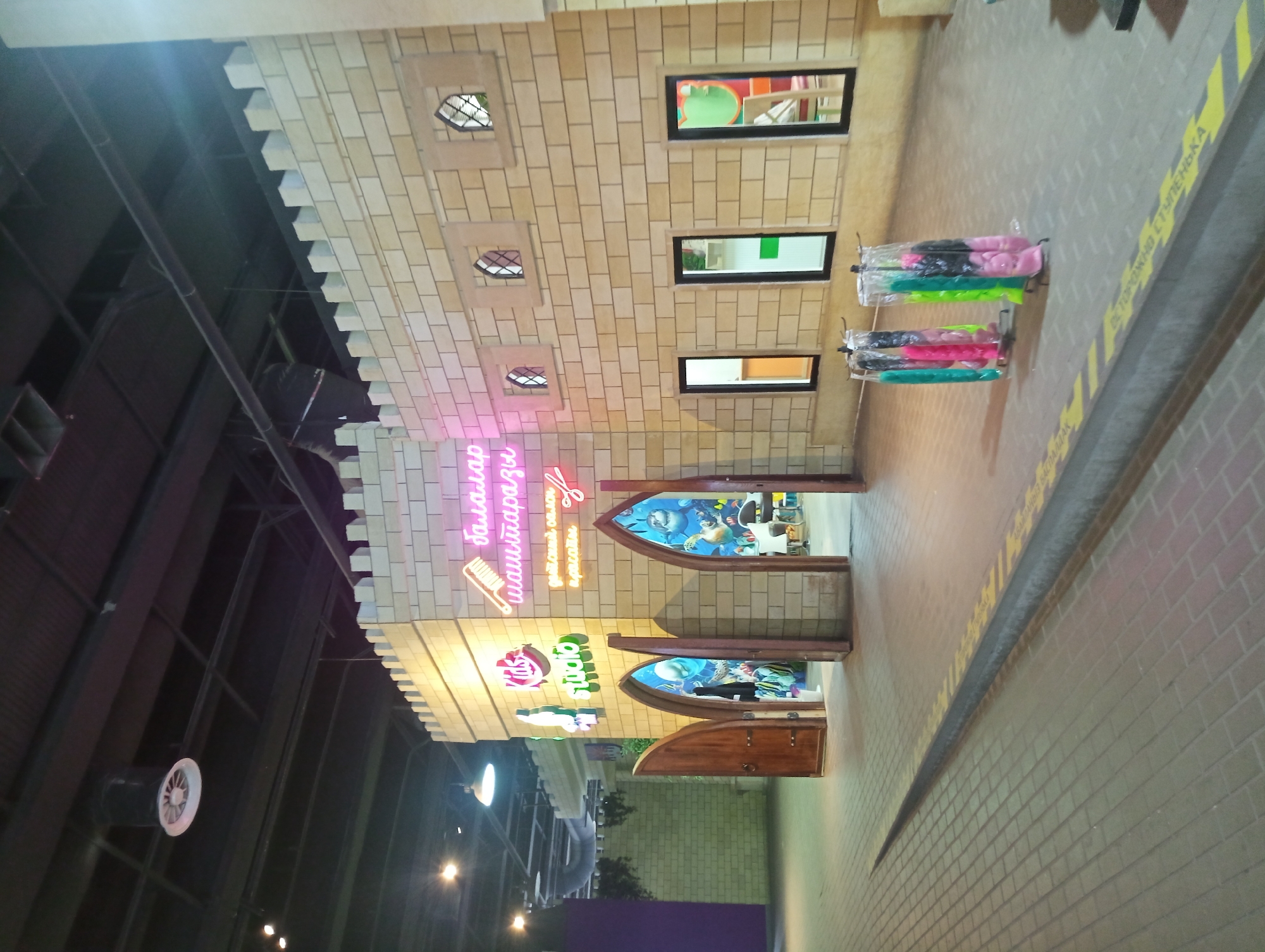 The image shows the exterior of a children's entertainment complex designed to resemble a castle. The facade is constructed from light-colored bricks and features crenellated battlements along the top. Several arched doorways, one open, reveal glimpses of the interior. Above one of the doorways, neon signs in pink and green spell out "Kids Studio" and other Cyrillic text. Various windows, some small and arched, others rectangular, are set into the wall. In front of the structure, several racks display colorful, individually wrapped items, possibly children's costumes or accessories, in shades of pink, green, and blue. The ground in front is paved with interlocking bricks and has a raised yellow and black striped border. The overall impression is that of a bright and engaging space for children's activities. The image does not contain enough visual information to pinpoint a specific location within the city of Almaty, Kazakhstan, or to identify any individuals.