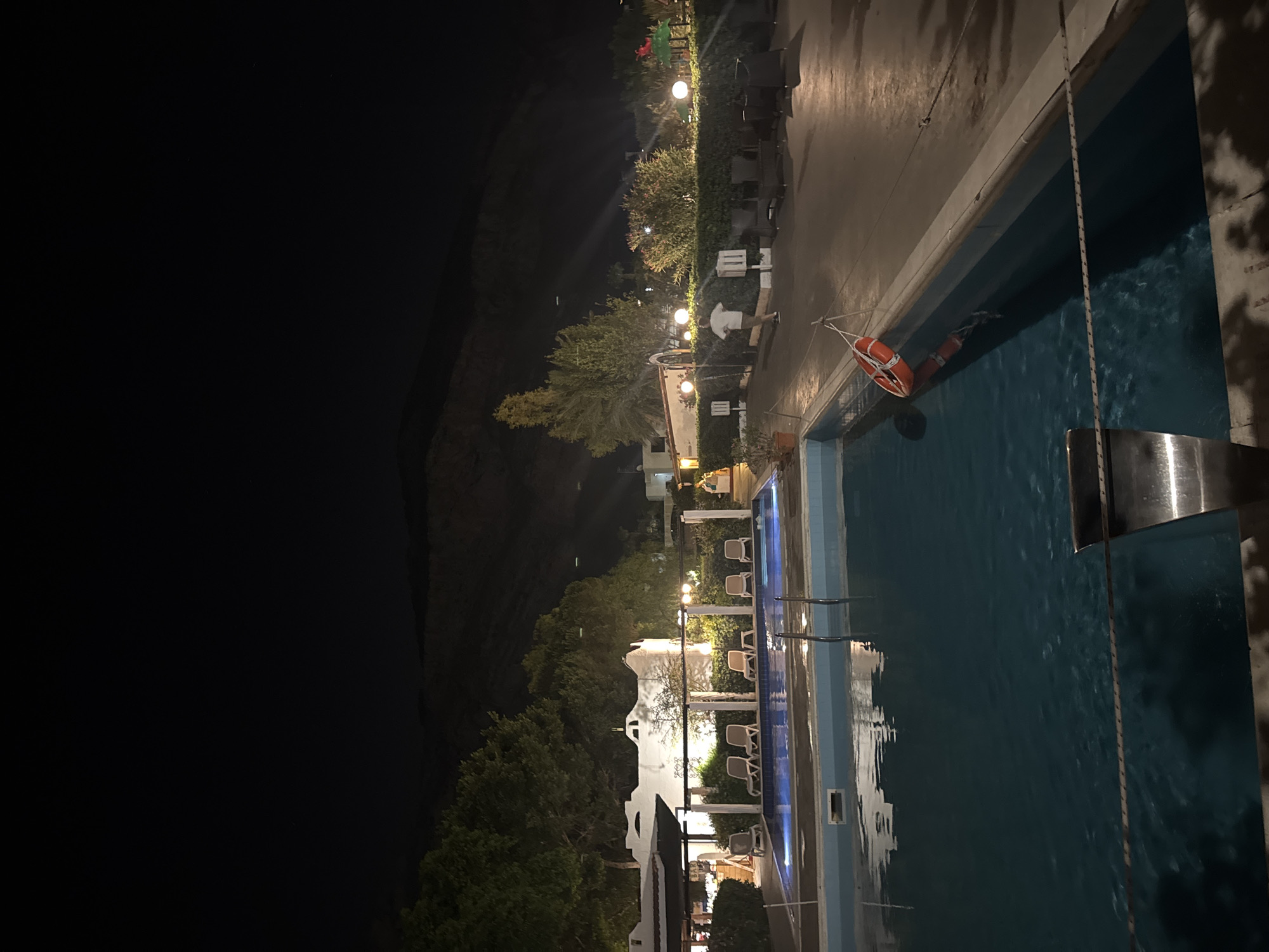 This is a nighttime, eye-level, medium shot of an outdoor swimming pool area. The pool is illuminated with underwater lights. To the left of the pool, there is a white building with an arched entryway and several lounge chairs arranged on a patio. Trees and bushes surround the building and patio. Further to the right, a person in a white t-shirt and light-colored shorts walks away from the camera on a paved pathway. There are several round, illuminated light fixtures along the pathway, and some seating areas with dark chairs and tables. A red life preserver ring hangs from a rope near the edge of the pool. In the background, a dark hillside is visible against the night sky. The overall scene suggests a resort or hotel environment. The location is identified as Isola delle Femmine, Italy.