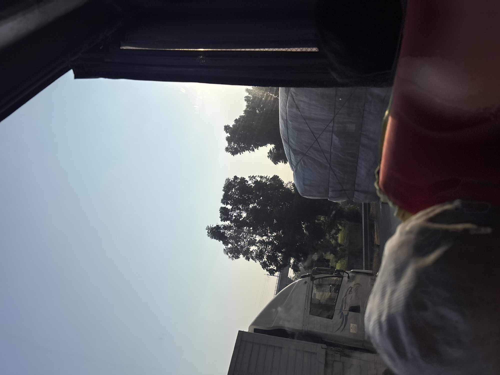 The image depicts a view from inside a vehicle, looking out at a road scene under a clear, pale blue sky. In the foreground, the heads of two passengers are visible, one with gray hair, and the other obscured by a red cushion. The window is framed by a dark curtain on the right. Outside, a white truck is partially visible on the left, its side and part of its cab showing. To the right of the truck, a large, tarp-covered load, likely on another vehicle, is prominent. Beyond these vehicles, trees with dense foliage stand against the sky, with a bridge or overpass visible in the distance. Sunlight streams through the trees, creating some lens flare and visible rays. The overall scene suggests a journey on a highway, possibly during the daytime given the lighting. The location is specified as the city of Hegarmukti, Indonesia.