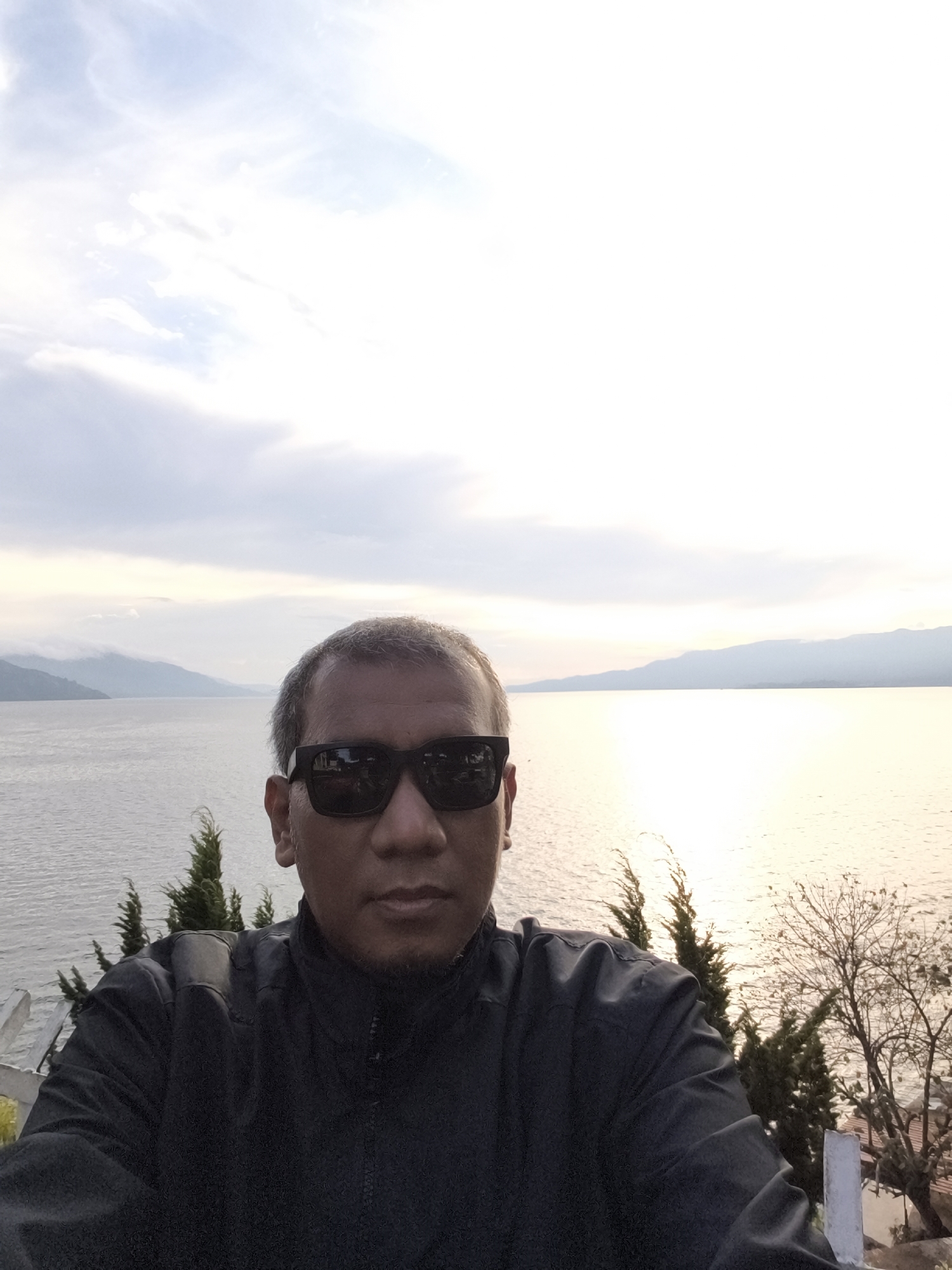 The image is a selfie of a man wearing sunglasses and a dark jacket, with a scenic background of a lake and mountains. The man is in the foreground, centered in the frame, with his face and upper body visible. He has short, graying hair and is looking directly at the camera. The background shows a wide expanse of water, likely Lake Toba, with rolling hills or mountains in the distance. The sky is partly cloudy, with the sun casting a warm glow on the water, suggesting it might be sunrise or sunset. The location is identified as Tiga Raja, Indonesia, which is situated on the shores of Lake Toba. The overall impression is one of a serene natural landscape, with the man posing for a photograph.
