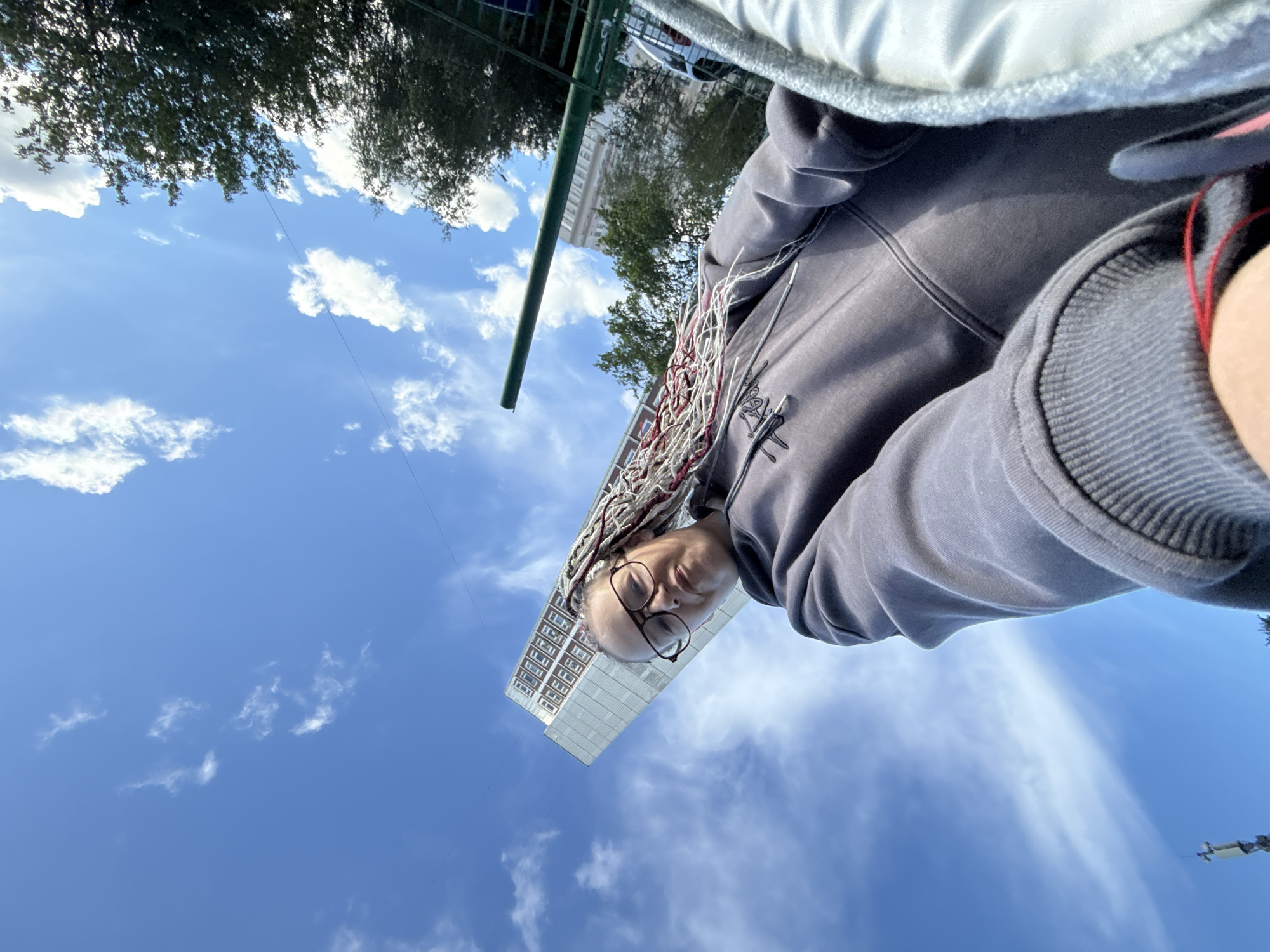 An upward-angle, first-person perspective selfie shows a woman with blonde and red braids wearing glasses and a gray hoodie. She is outdoors with a bright blue sky and scattered white clouds above. A tall, modern apartment building with many windows is visible in the background, along with trees and a green fence. A cellular tower is also visible in the distance against the sky.