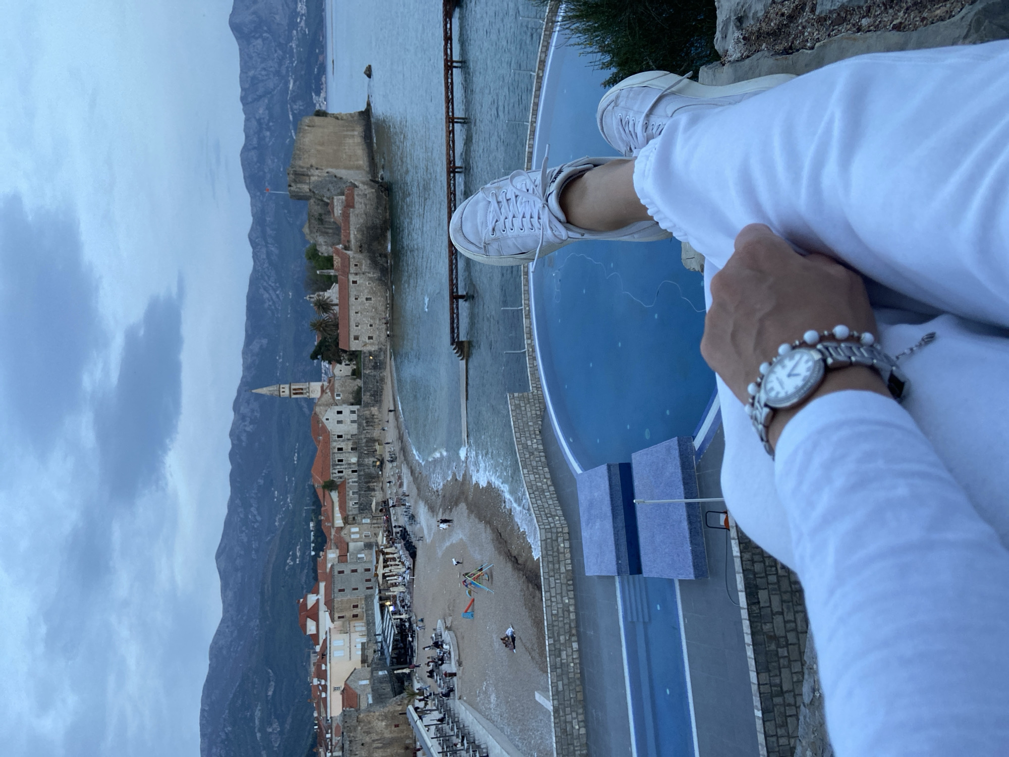 A person wearing white clothing and white sneakers sits on a ledge, overlooking a coastal town with mountains in the background. The town has buildings with red-tiled roofs, a beach with people and objects, and a historic fortress. A pool is visible below the ledge. The person is wearing a watch and bracelet on their left wrist. The photo is taken from a high vantage point. The town is the old town of Budva, Montenegro.
