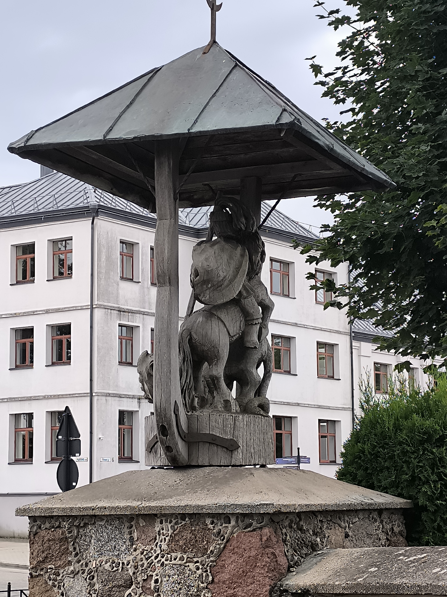 The image shows a wooden sculpture of a knight on horseback, possibly a representation of a historical figure or a local legend. The sculpture is weathered and appears to be quite old, possibly dating back to the mid-20th century. The knight is depicted with a shield and what appears to be a sword, although the details are obscured by the material and the lighting. The sculpture is sheltered under a dark, metallic roof supported by wooden beams. Below the sculpture, there is a stone pedestal constructed from a mosaic of various-sized stones, including reddish-brown and greyish varieties, interspersed with mortar.
The sculpture and its base are positioned in front of a white, multi-story building with a metal-plated roof. The building has numerous windows with dark frames. To the right of the building, there is dense green foliage, likely a tree or shrub. In the foreground, there are street signs, one triangular and black, and another rectangular with white lettering, indicating street names. A glimpse of a black metal fence is visible at the bottom left of the frame.
This monument is located in the town of Kretinga, Lithuania, specifically in the vicinity of the Kretinga Regional Museum, which houses a significant collection of historical artifacts and art. The building in the background is likely part of the museum complex or a nearby public institution. The overall atmosphere suggests a public space, possibly a courtyard or a small square within the town. The sky is overcast, contributing to the muted color palette of the scene.