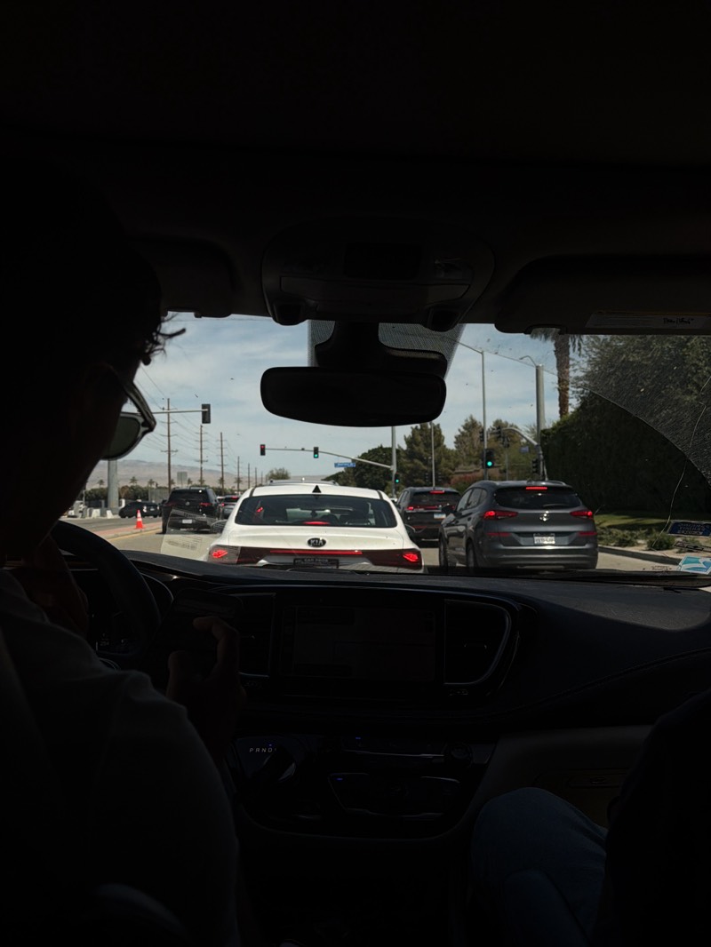 The image depicts a view from inside a car in La Quinta, United States. The driver, wearing sunglasses, is behind the wheel of a Chrysler Pacifica with a black dashboard. Outside the windshield, there's a white Kia and a gray Hyundai Tucson, among other cars stopped at a traffic light on a street with telephone poles and palm trees. The sky is clear and lightly clouded.