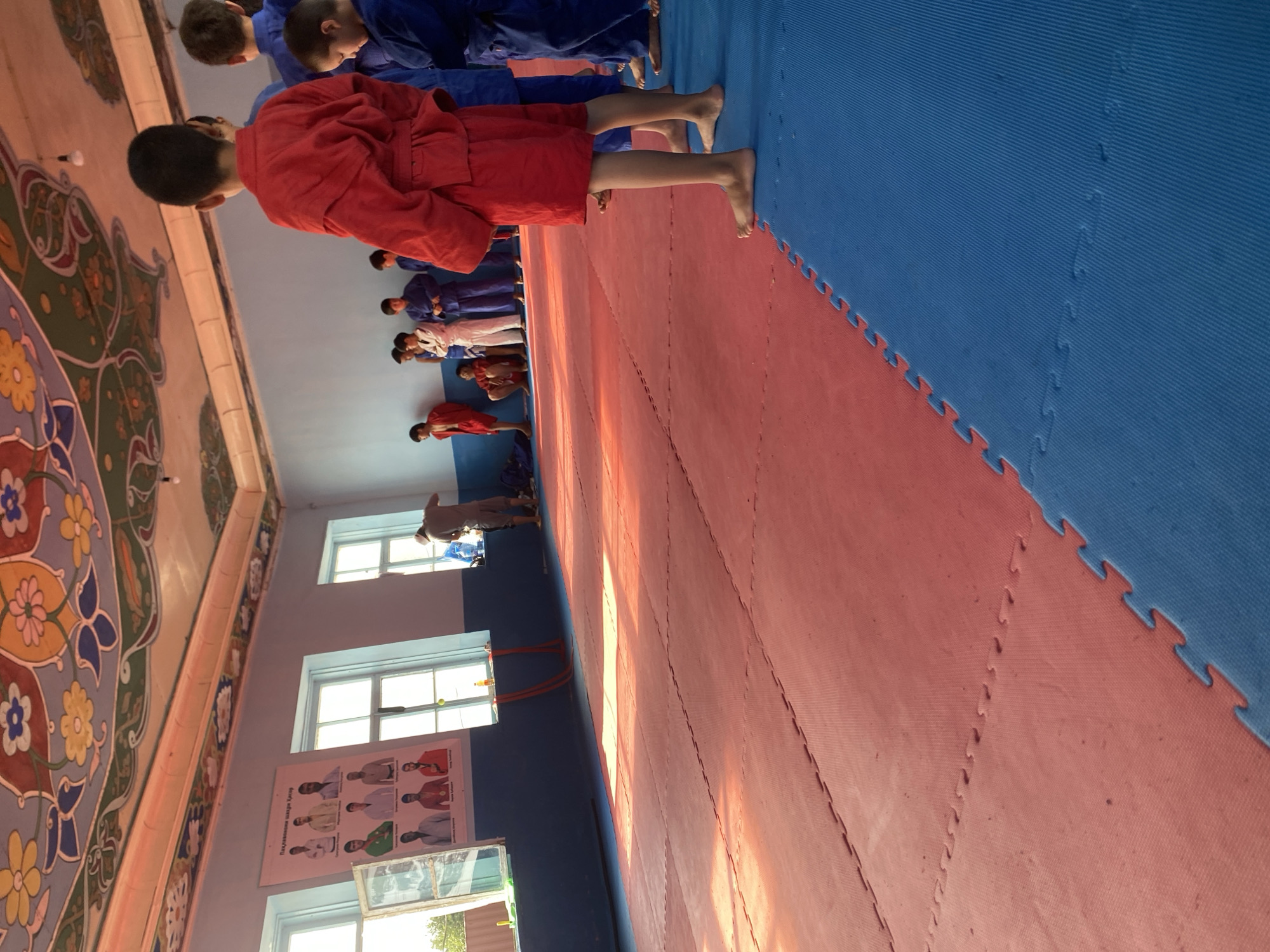 The image shows the interior of a sports hall, likely a judo or sambo gym, in Боғистон, Tajikistan. The hall has a distinctive, brightly colored, floral-patterned ceiling. The floor is covered with interlocking puzzle mats, divided into red and blue sections.
In the foreground, a group of children and young people are gathered. Most are wearing traditional martial arts uniforms, with some in blue and one in a red uniform prominently displayed in the right foreground. They are facing towards the left of the frame, suggesting they are either waiting for instruction or observing an activity.
On the left side of the hall, near a window, there is a poster on the wall displaying portraits of several individuals, some in martial arts attire and others wearing medals. The poster appears to be educational or motivational, possibly showcasing champions or instructors.
The environment is bright, with sunlight streaming through the windows, casting long shadows across the red mats. In the background, near another window, a person in a grey shirt and shorts is attending to something, possibly preparing equipment or cleaning. The overall scene depicts a training session or a gathering in a martial arts facility.