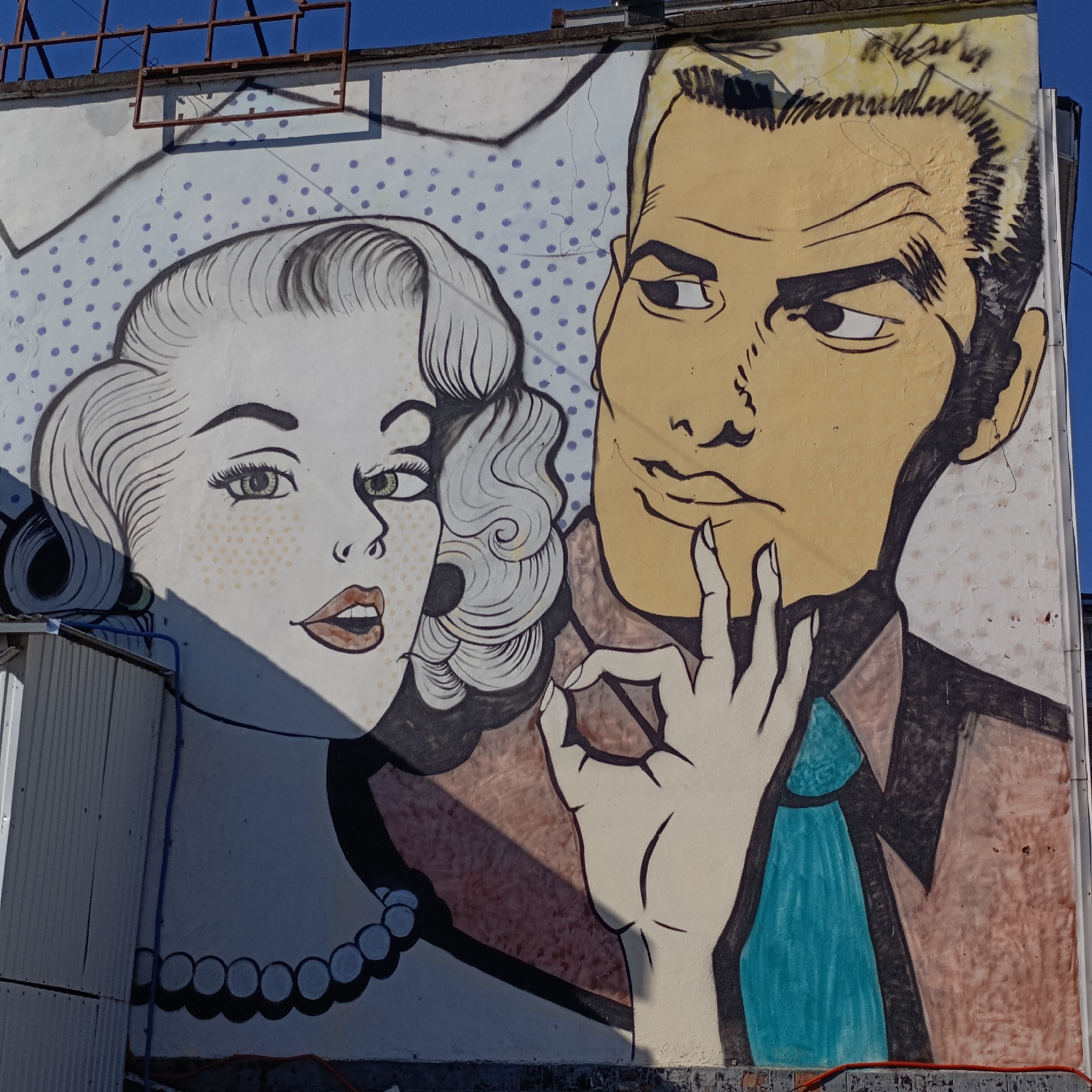 The image is a mural depicting a man and woman in a cartoon style reminiscent of mid-20th century comic books. The woman, on the left, has light skin, green eyes, and her hair styled in waves. She wears a pearl necklace and has a surprised expression. The man, on the right, has blond hair, tan skin, and a pensive expression with his fingers touching his chin. He is wearing a collared shirt with a tie. The background behind them is a light-colored wall with a pattern of dots. The mural is located on the side of a building in the city of Chaikovsky, Russia. Above the mural is a metal framework, and to the left is a metal structure. The image appears to be taken during daylight under clear skies. The bottom of the mural is partially obstructed by a small wall or curb.