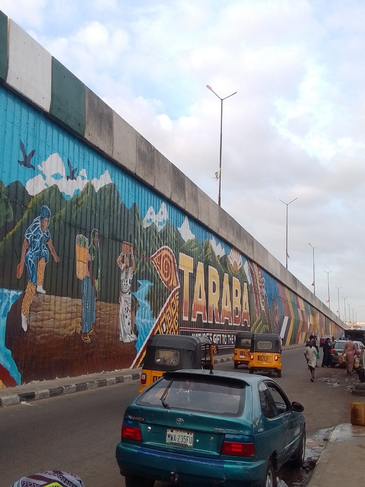 The image depicts a wide street in Jalingo, Nigeria, dominated by a large, colorful mural on a concrete overpass. The mural features scenes of people, mountains, birds, and abstract geometric patterns. The overpass itself has sections of white, green, and grey concrete. Streetlights are visible along the overpass, with a few flags attached to poles.
On the street, several vehicles are present. A teal Toyota Corolla is parked in the foreground, facing away from the camera, with a Nigerian license plate that reads "MWA 235FU". Two yellow auto-rickshaws, also known as "keke napep", are driving on the road. One has a license plate "BD21 U" and the other has "1020 U". Further down the street, other vehicles, including cars and more auto-rickshaws, are visible, along with several people walking. The environment appears to be a busy urban setting with cloudy skies.