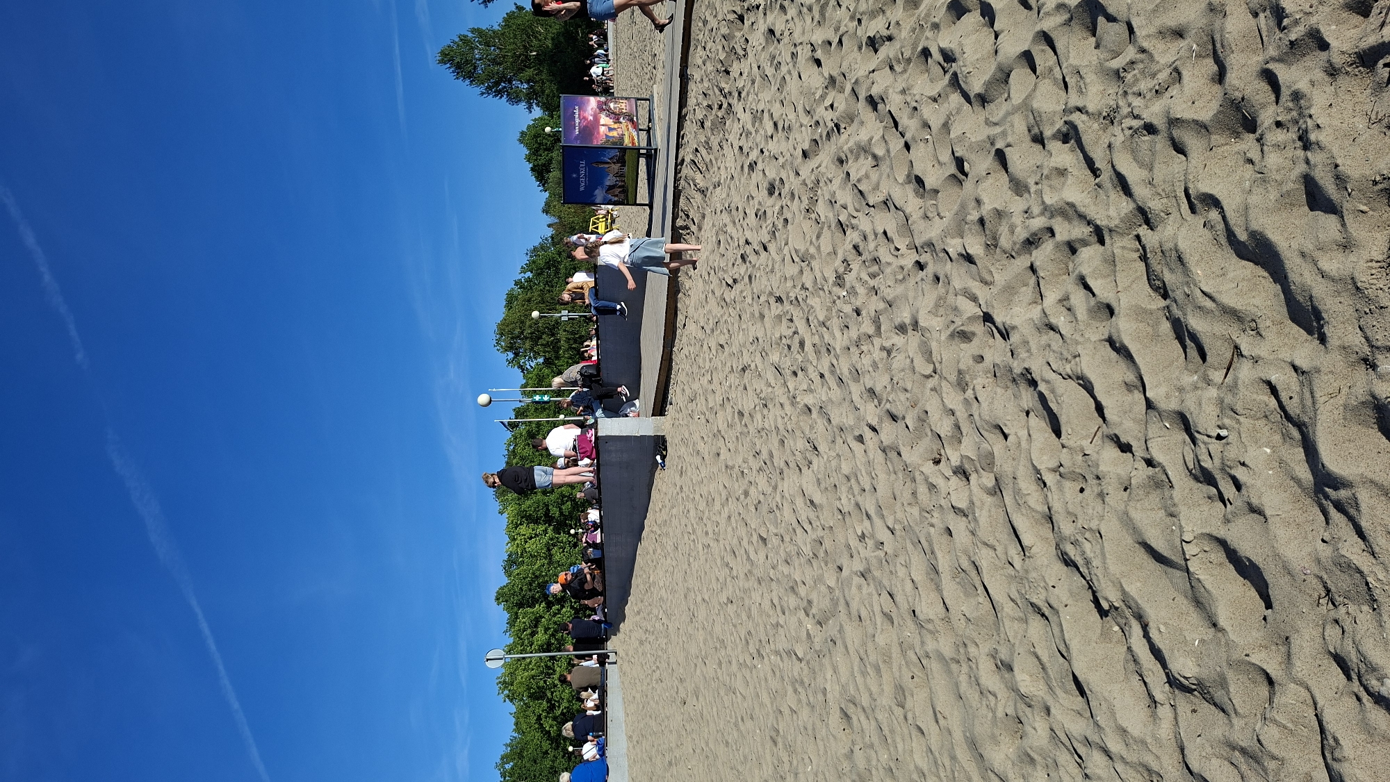 The image shows a sunny day at a sandy beach in Tallinn, Estonia, likely during the summer. The foreground is dominated by rippled sand, with scattered small debris. In the midground, a group of people are seated and standing along a raised concrete wall, behind which a line of trees forms a natural backdrop against a clear blue sky. A girl in a light-colored skirt and top is walking on the sand in the midground, facing away from the camera. Further in the background, there are promotional displays and more people. The lighting suggests it is late afternoon or early evening, with long shadows cast by the undulations in the sand. The presence of trees and the general atmosphere indicate a recreational area within the city.
