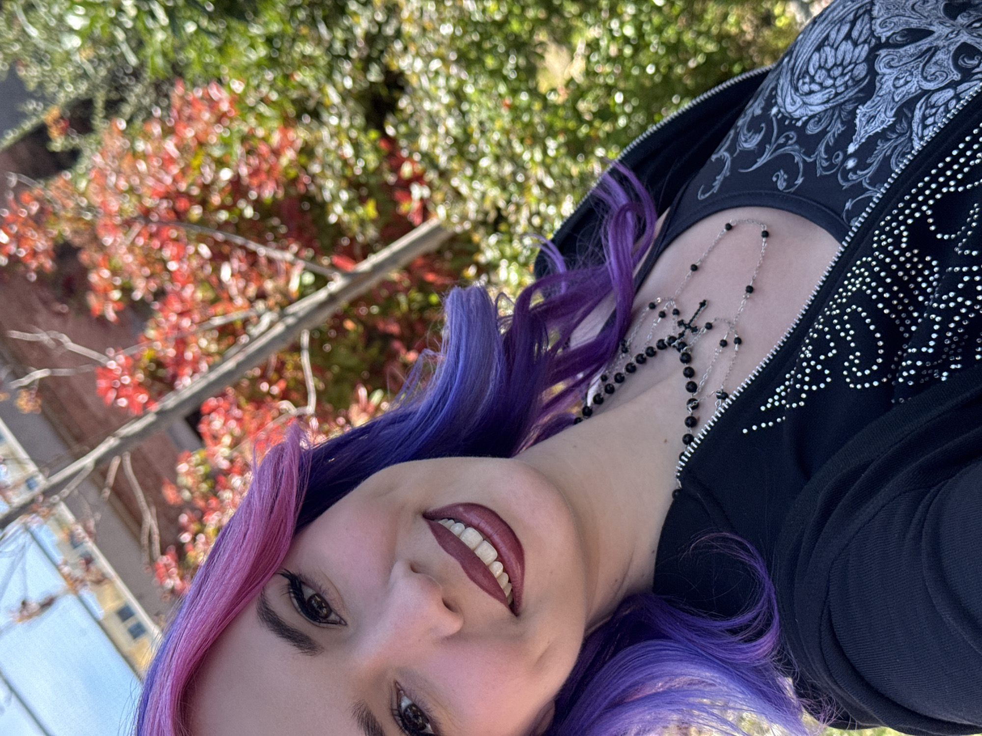 A woman with purple and pink hair is seen in a close-up, tilted shot. She is smiling, wearing dark lipstick and a black outfit. Around her neck is a silver chain with black beads and a cross. The background shows trees with red and green foliage. Based on the architecture and foliage visible, the image may be taken in a park-like setting in Madrid, Spain.