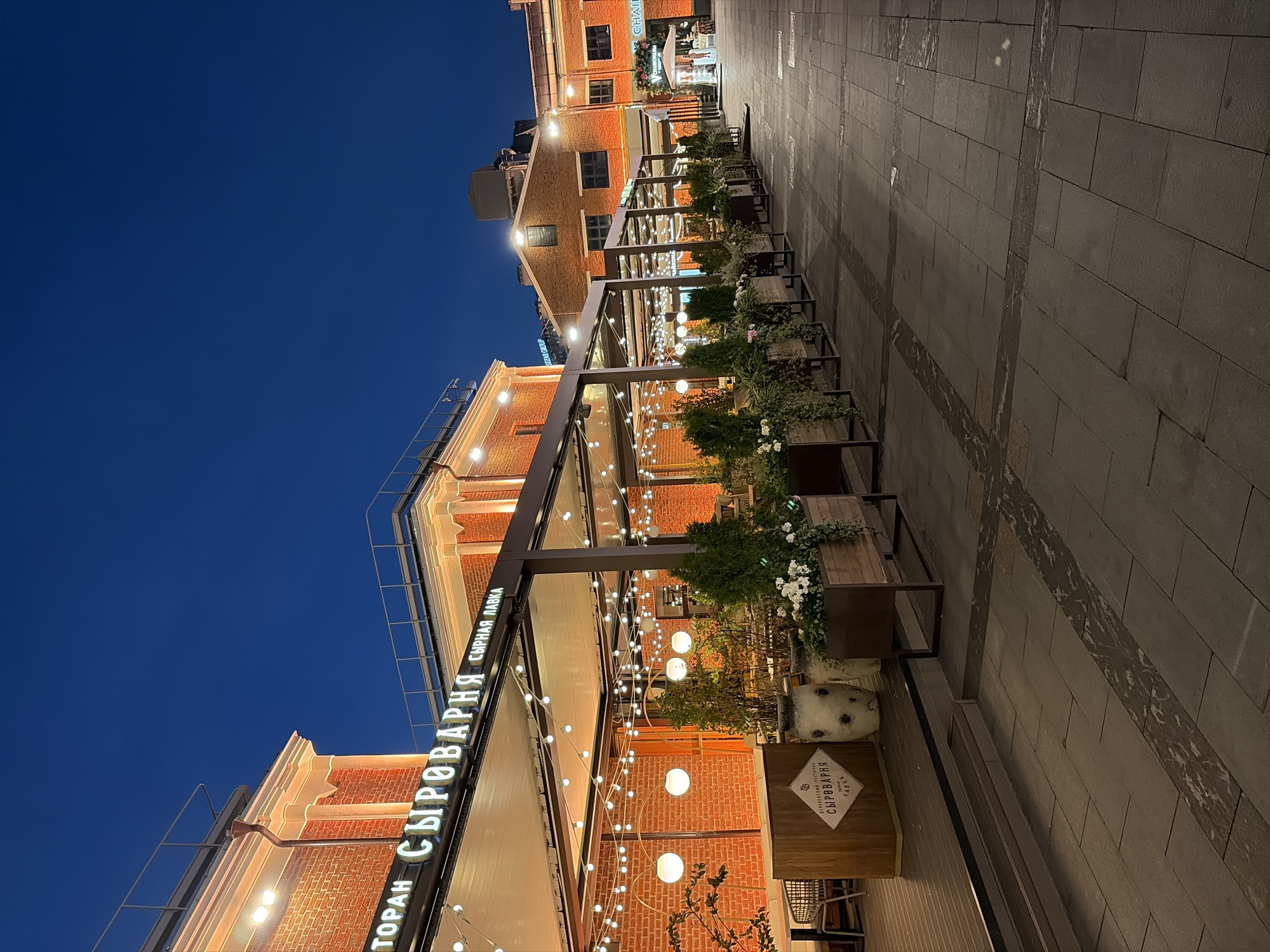 The image captures a nighttime view of the outdoor seating area of "SYROVARNYA" (СЫРОВАРНЯ), a restaurant in Moscow, Russia, likely located at a former industrial site repurposed into a modern urban space. The facade of the building is made of red brick, illuminated by warm interior lights and decorative string lights. The restaurant's name is displayed in prominent white neon letters above the entrance.
The outdoor seating area is covered by a retractable canopy, also adorned with numerous string lights that create a festive and inviting atmosphere. Several tables and chairs are visible, some with a modern, minimalist design. Planters filled with greenery and flowers line the walkway in front of the restaurant, adding a touch of nature to the urban setting. Large, decorative ceramic pots also contribute to the ambiance.
The ground is paved with large, rectangular stone tiles, with a decorative pattern of darker stone interspersed. The sky above is a deep, dark blue, indicating it is nighttime or twilight. In the background, other buildings with brick facades and illuminated windows are visible, suggesting this is part of a larger complex or development, common in Moscow's revitalized industrial districts. No people are clearly visible in the frame, but the setup suggests an active dining environment. The overall impression is one of a sophisticated and comfortable urban dining experience.
