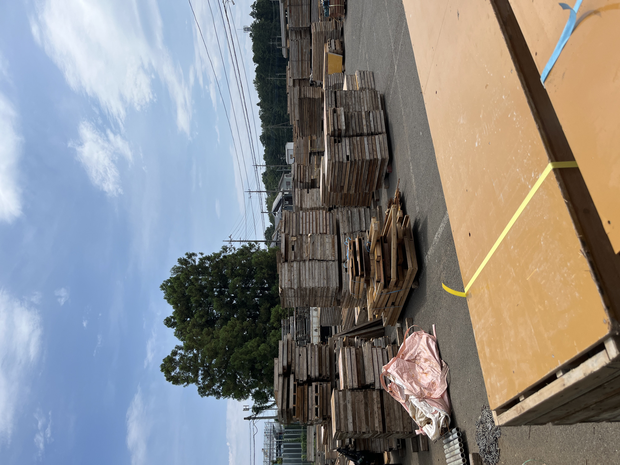 An outdoor, daylight scene in Sayama, Japan, captures a storage area filled with stacked wooden planks and building materials under a partly cloudy sky. The foreground features a large, flat, orange-brown surface with a yellow strap and a blue strap. To the left, a pile of metal chain is visible, next to a stack of cylindrical metal pipes. Further into the scene, numerous stacks of wooden planks of varying sizes are arranged in neat piles. A large, pinkish fabric bag is also present. In the background, a truck is partially visible, and dense green trees form a backdrop. Power lines crisscross the sky. The ground appears to be asphalt or a similar paved surface. No people are visible in the image.