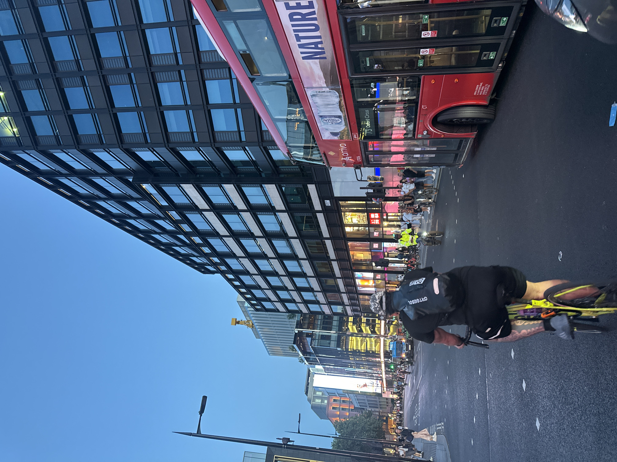 The image depicts a street scene in London, United Kingdom, likely during dusk. A red double-decker bus, advertising "NATURE" and a bottle of water, is prominent on the right side of the frame. In the foreground, a person wearing a helmet and a black backpack is cycling away from the camera, their legs and tattoos visible. The street is paved with asphalt and marked with white lane lines.
On the left side of the street, there are several multi-story buildings with modern architecture, featuring large glass windows and illuminated signs. Pedestrians are visible walking on the sidewalks and some are crossing the street. A traffic light is illuminated green for the vehicles. Further down the street, more buildings and streetlights are visible. Another cyclist, wearing a high-visibility jacket, is seen in the middle ground, also on a bicycle. The sky is a clear, twilight blue. The overall impression is a busy urban environment transitioning into evening.