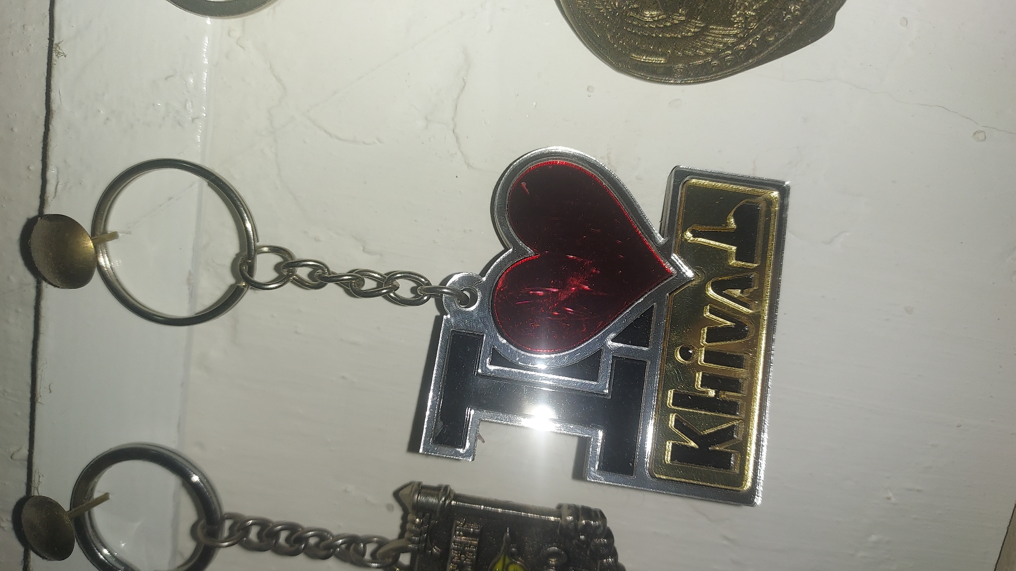 This is a close-up, eye-level, indoor photograph of several keychains hanging from a white wall. The keychains are attached to the wall with small, round, brass-colored fasteners. The main subject is a silver and black keychain with a red heart and the word "Khiva" in gold lettering. To the left of this keychain is another keychain with a silver-colored building. To the right, a portion of a gold-colored coin or medallion is visible. The wall appears to be painted white and has some minor imperfections and shadows. There are no people visible in the image. This image is not relevant to the city of Kogon City, Uzbekistan, as the keychain clearly indicates the city of Khiva.