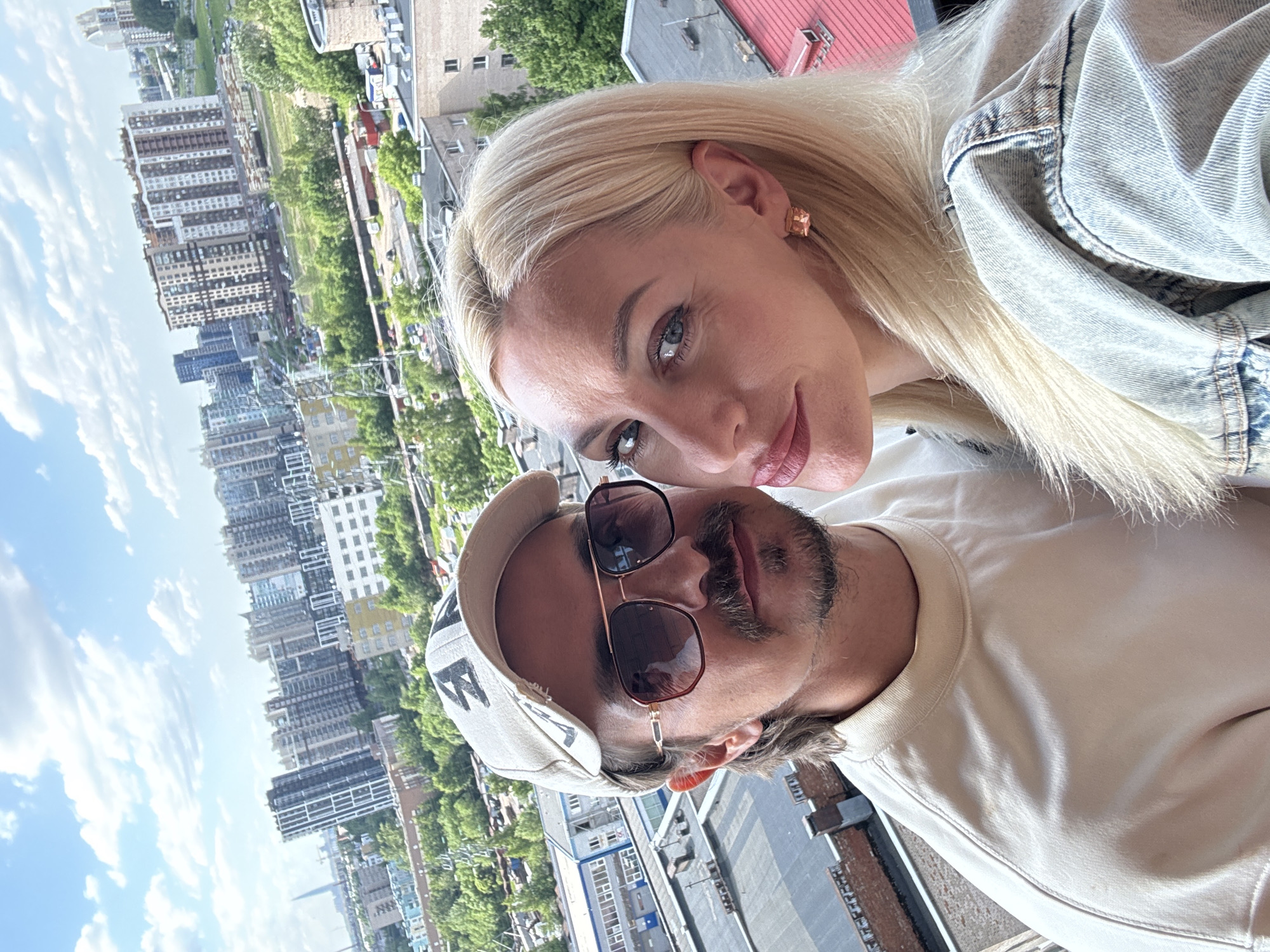 This is a medium shot selfie of a couple in front of an urban landscape. The man on the left is wearing a light-colored baseball cap, sunglasses, and a light-colored crewneck sweatshirt. He has a mustache and a goatee, and his hair is visible below the cap. The woman on the right has blonde hair and is wearing a light-wash denim jacket and a square-shaped earring. They are positioned close together, with the woman leaning in towards the man. The background shows a densely populated city with numerous apartment buildings of varying heights and architectural styles, interspersed with green trees. A cloudy sky with blue patches is visible above the buildings. The scene appears to be an outdoor setting, possibly a balcony or rooftop, given the elevated perspective of the city.