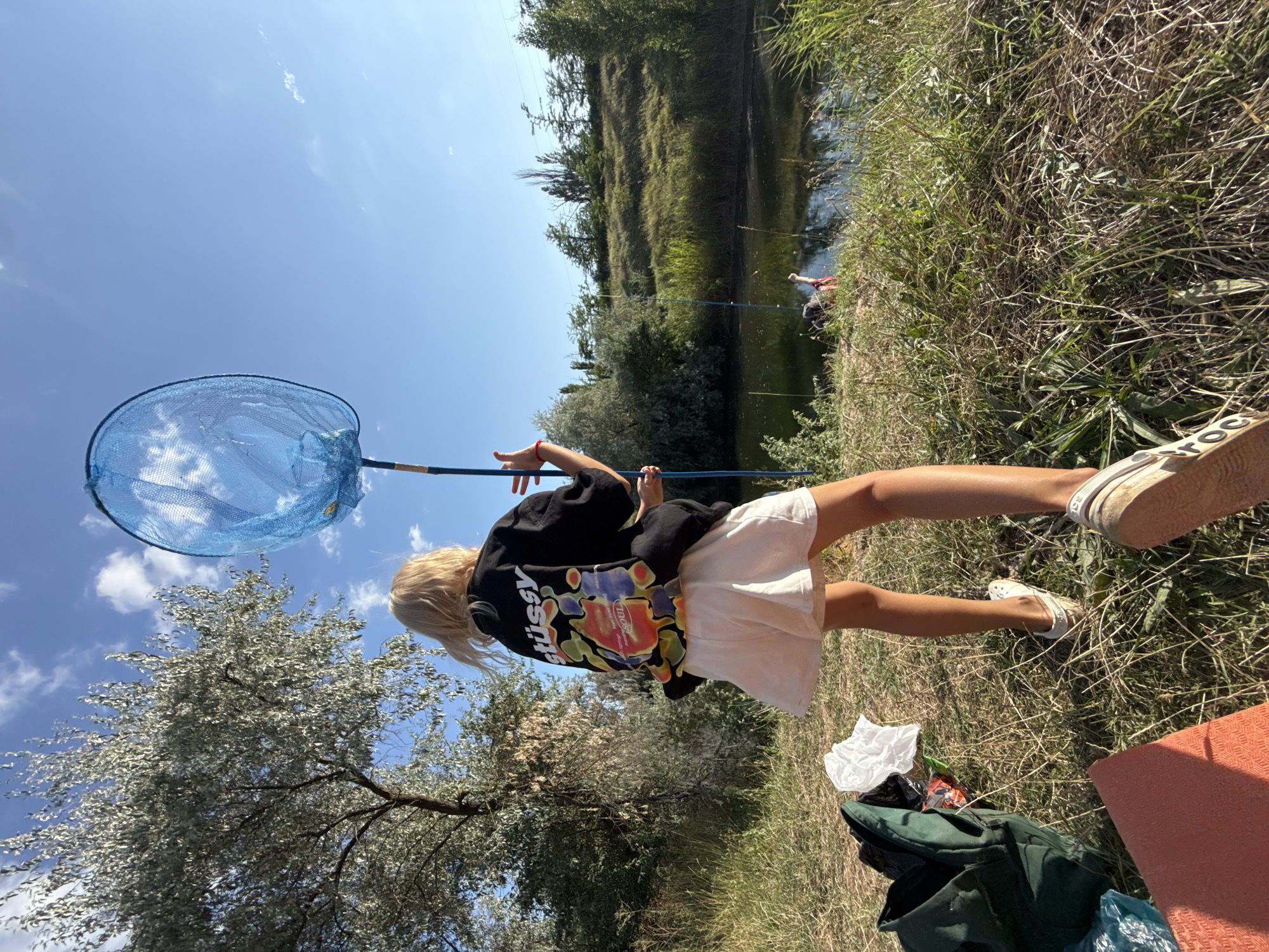 A young girl with blonde hair stands in a grassy field, holding a blue fishing net above her head. She is wearing a black t-shirt with a colorful design on the back and a white skirt. In the background, there is a body of water with trees and a clear blue sky with some clouds. There are two fishing rods in the water, and a person in the water wearing a red shirt. A green backpack and an orange mat are on the ground near the girl. The scene appears to be a rural area, possibly on the outskirts of Пушкинское сельское поселение, Ukraine.