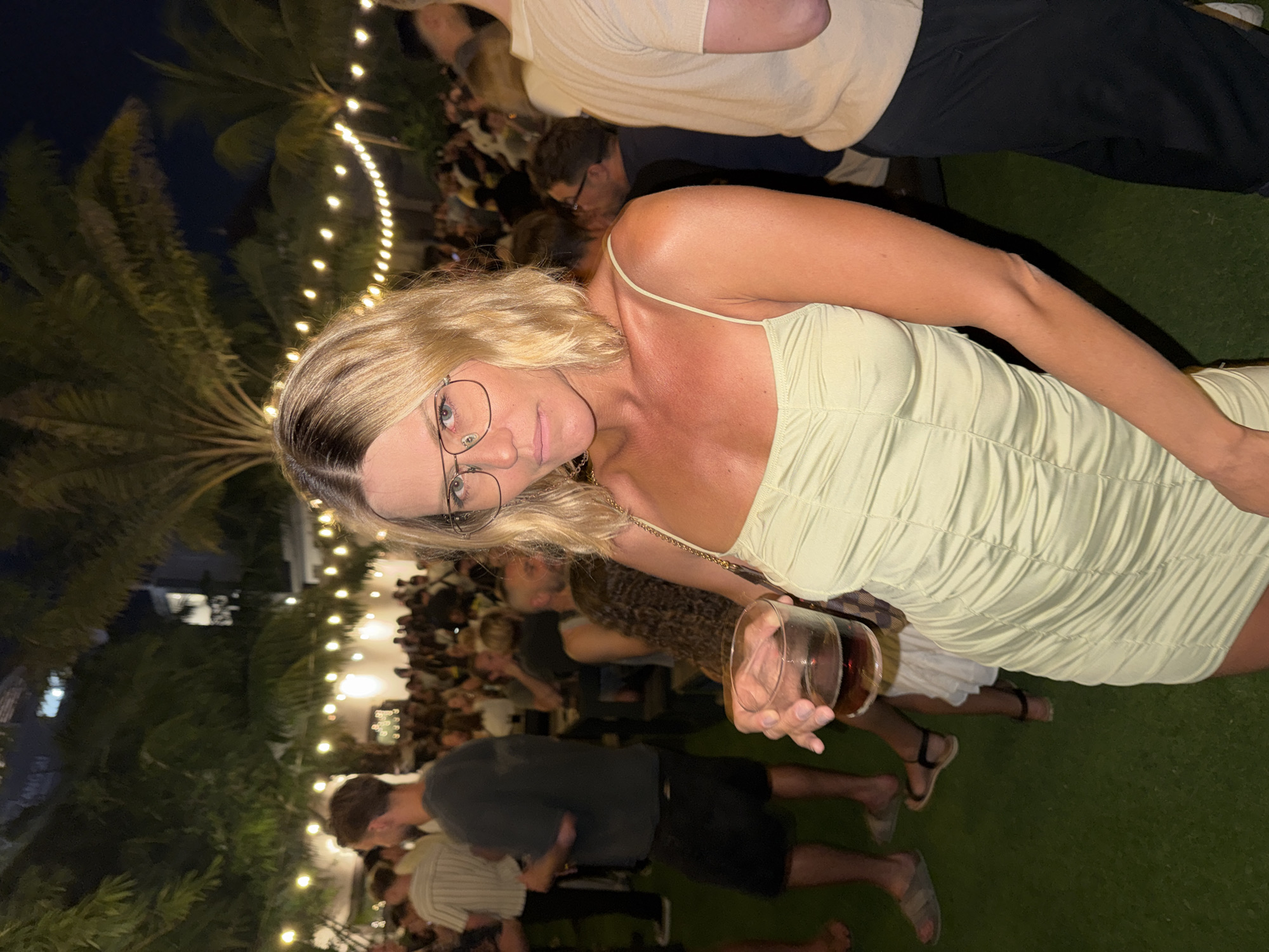 The image shows a blonde woman in a light green dress holding a glass of dark liquid in what appears to be an outdoor event with many people in the background. The event seems to be taking place at night, as indicated by the presence of strung lights and palm trees. The photograph was taken in North Kuta, Indonesia.