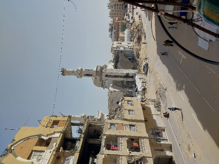 The image depicts a street scene in Khan Yunis, Palestinian Territory, characterized by extensive structural destruction under a clear, bright sky. In the mid-ground left, a multi-story building, appearing residential, shows severe damage with entire sections of its facade and internal structures collapsed, revealing exposed rooms. Laundry hangs on remaining balconies. In the background center, a large mosque with a white dome and a tall, slender minaret stands heavily damaged, with significant portions of its structure crumbled or missing.
The street itself is a paved road with faded white lane markings. Rubble and debris are scattered along the sides of the road and in open areas adjacent to the damaged buildings. Numerous individuals are present on the street level; some walk alone, while others are in small groups. One person in a striped shirt walks in the foreground street. A light-colored sedan is visible parked amidst the rubble in the mid-ground. The scene is illuminated by strong sunlight, casting long shadows from buildings and individuals onto the pavement. The overall environment appears dusty and affected by widespread damage. The foreground right is partially obscured by a dark, curved metal object, possibly a railing.
