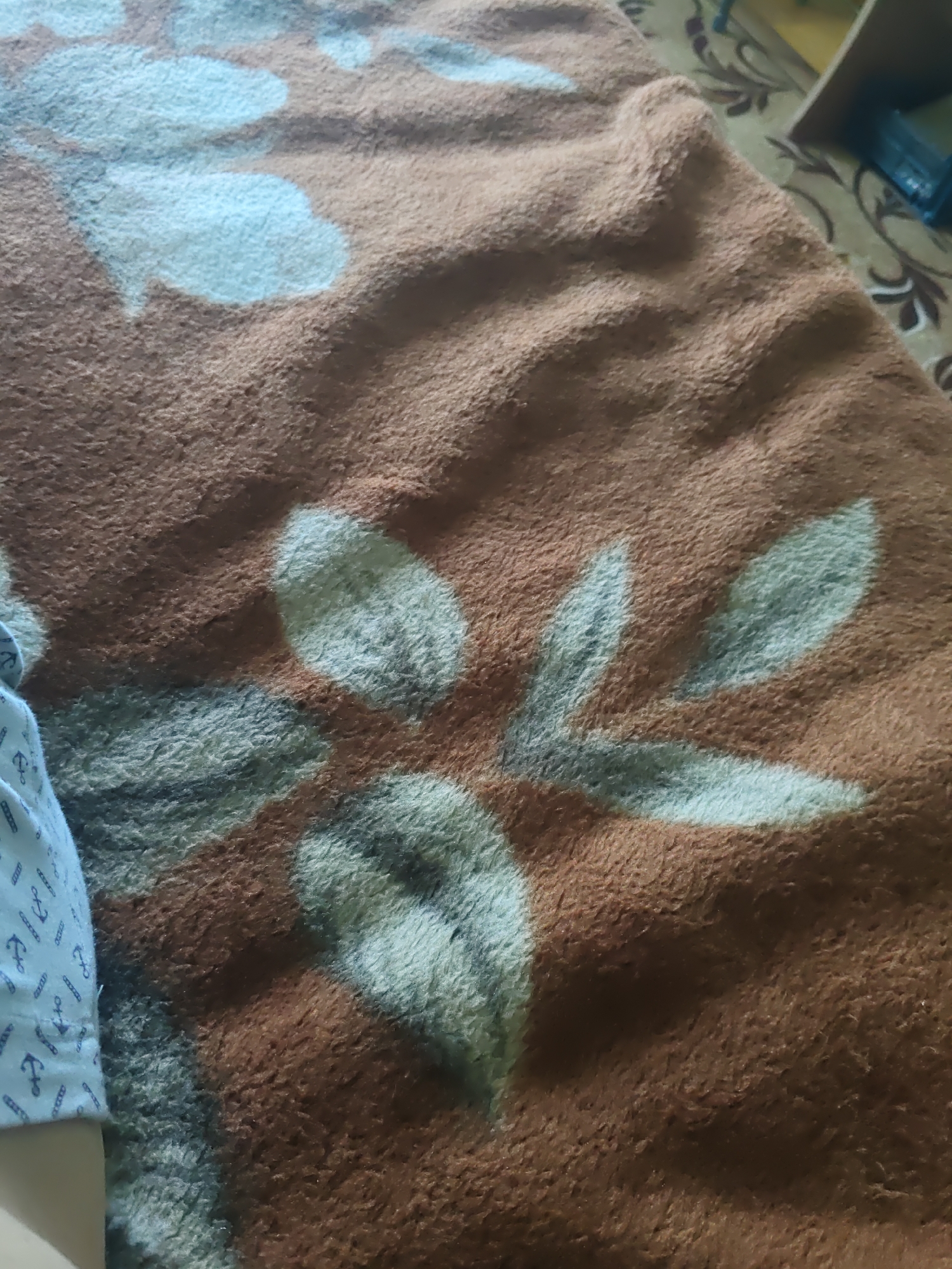 The image shows a close-up of a brown blanket with a light blue floral pattern. The blanket is textured and appears to be made of a soft material. On the left side of the image, a portion of a light blue shirt with a pattern of dark blue anchors is visible. The shirt is worn by an unseen person. The background of the image is out of focus, but it appears to be a patterned carpet or rug. There are no discernible elements that would indicate the specific location within the city of Курашим, Russia.