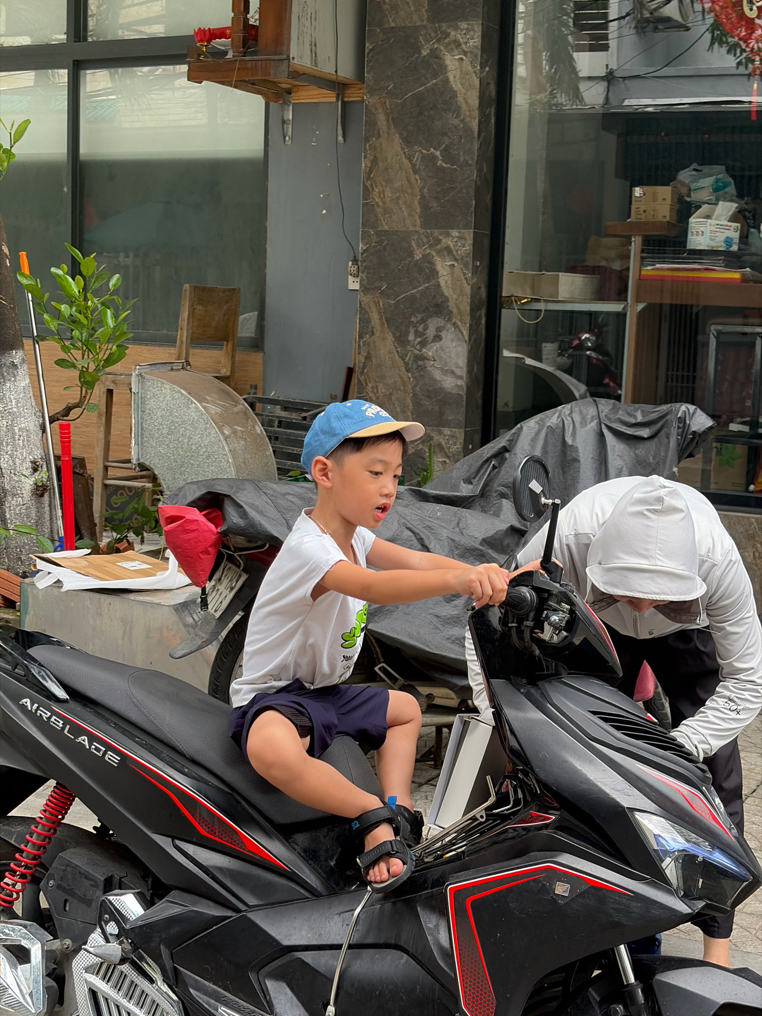 In Đà Nẵng, Vietnam, a young boy wearing a blue baseball cap and a white t-shirt sits on a black and red motorcycle. He is looking to the right and appears to be interacting with a person behind him, who is wearing a white hoodie and black pants. The person's face is obscured by a mask. The motorcycle is parked on a paved surface in front of a building with a large window and marble-like facade. Various objects are scattered around, including a red pole, a plant, and some cardboard boxes. A tarp is draped over part of the motorcycle and a nearby structure. The scene suggests a casual outdoor environment, possibly a street or a yard, in an urban setting.