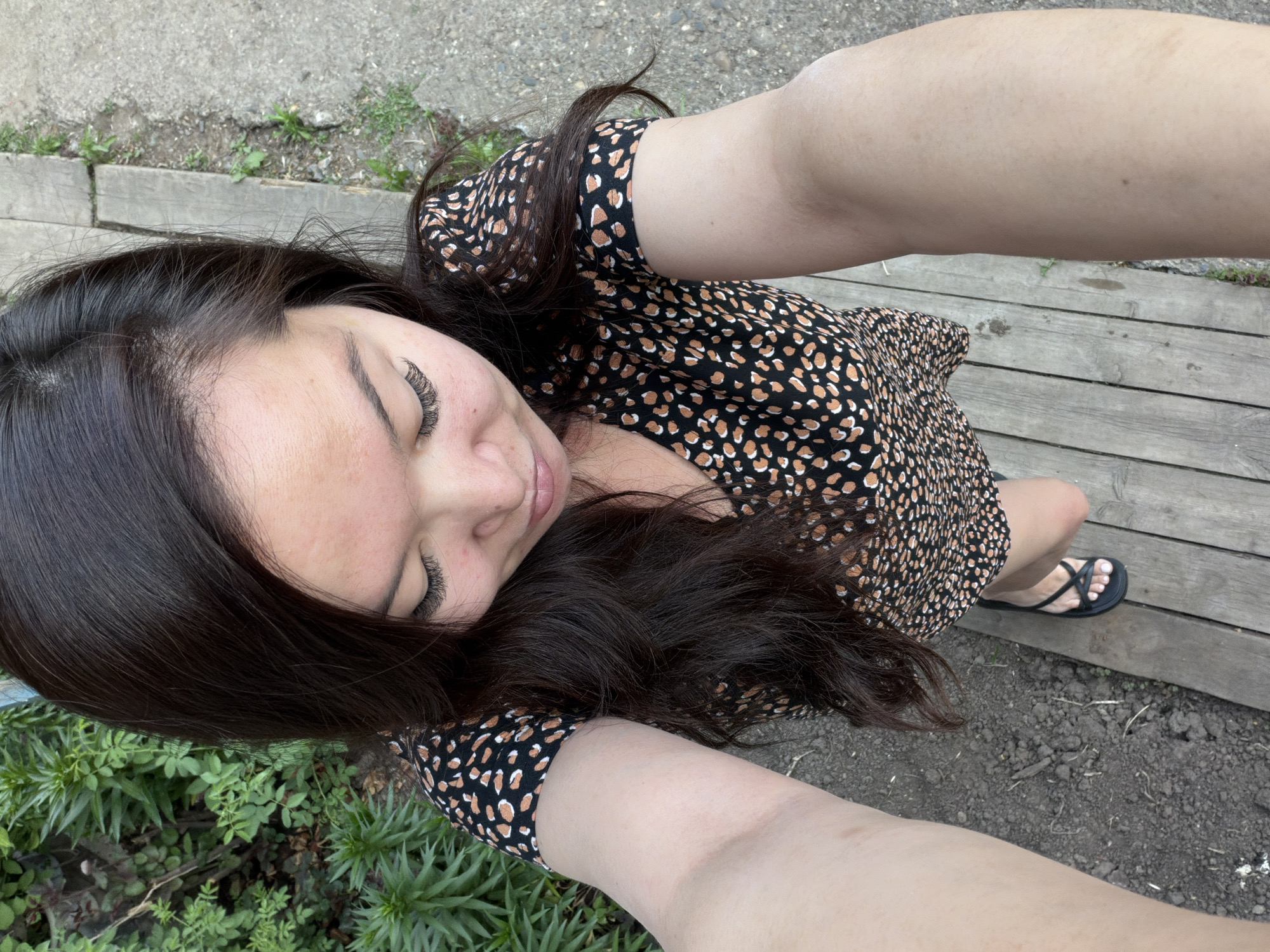 A woman with dark hair and false eyelashes is taking a selfie outdoors. She is wearing a black and tan animal print dress and black sandals. She is standing on a wooden deck or platform with grass and plants visible around it. Concrete and dirt are also present in the surrounding area. The location is unspecified; no information is available to determine if the photograph was taken in the city of Izhevsk, Russia.