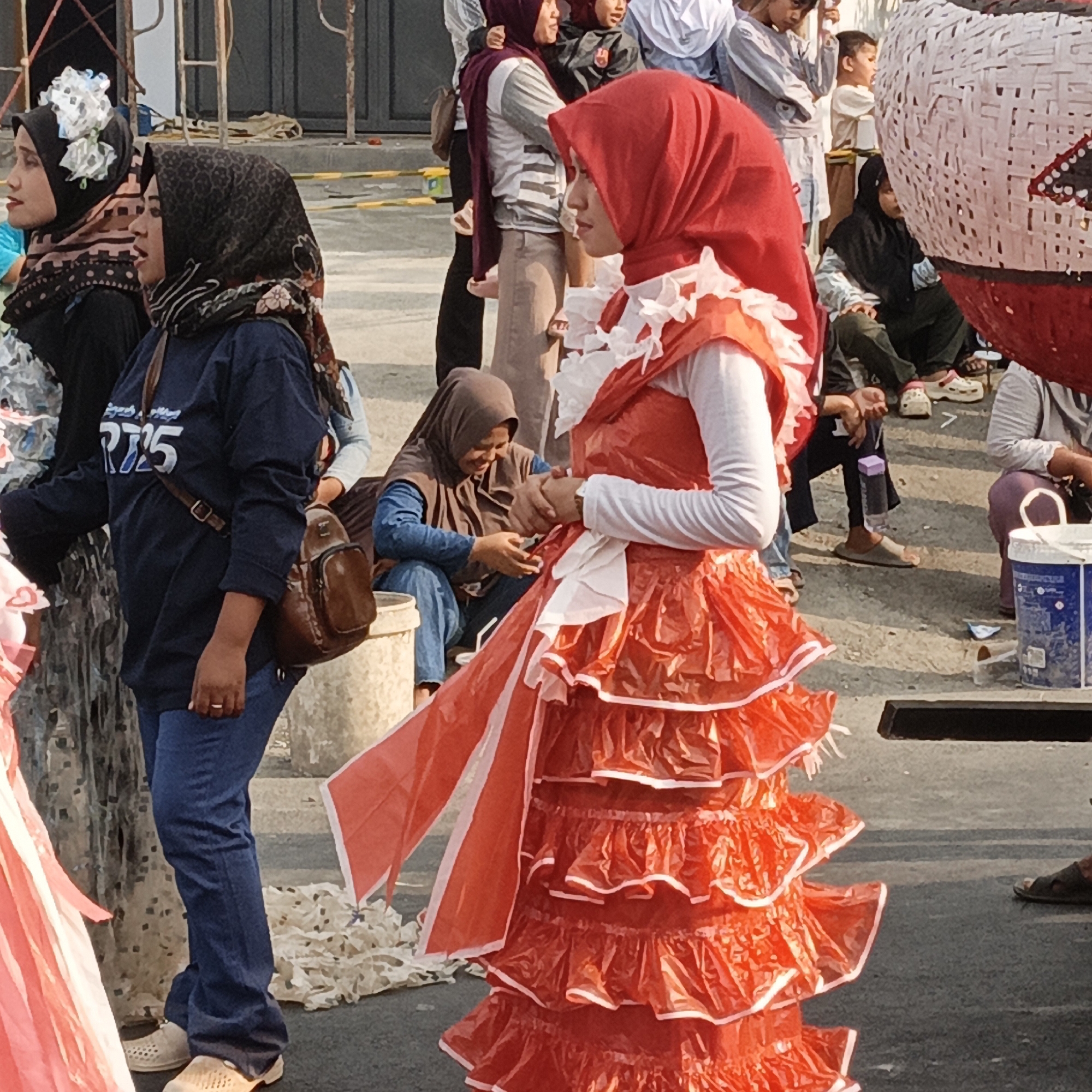 A group of people, primarily women and children, are gathered outdoors in what appears to be an urban environment. The scene is characterized by the presence of various garments, including hijabs and traditional clothing. One woman in the foreground is wearing a vibrant orange dress with ruffled layers, possibly a costume. She is adorned with a red hijab. Other individuals are seen in the background, some are seated, while others stand. Objects such as buckets and bags are scattered around. The ground surface is paved, and there are signs of construction or maintenance work in the background, with metal scaffolding and barriers visible. The lighting suggests it is daytime. The overall atmosphere seems to be one of a community event or gathering.