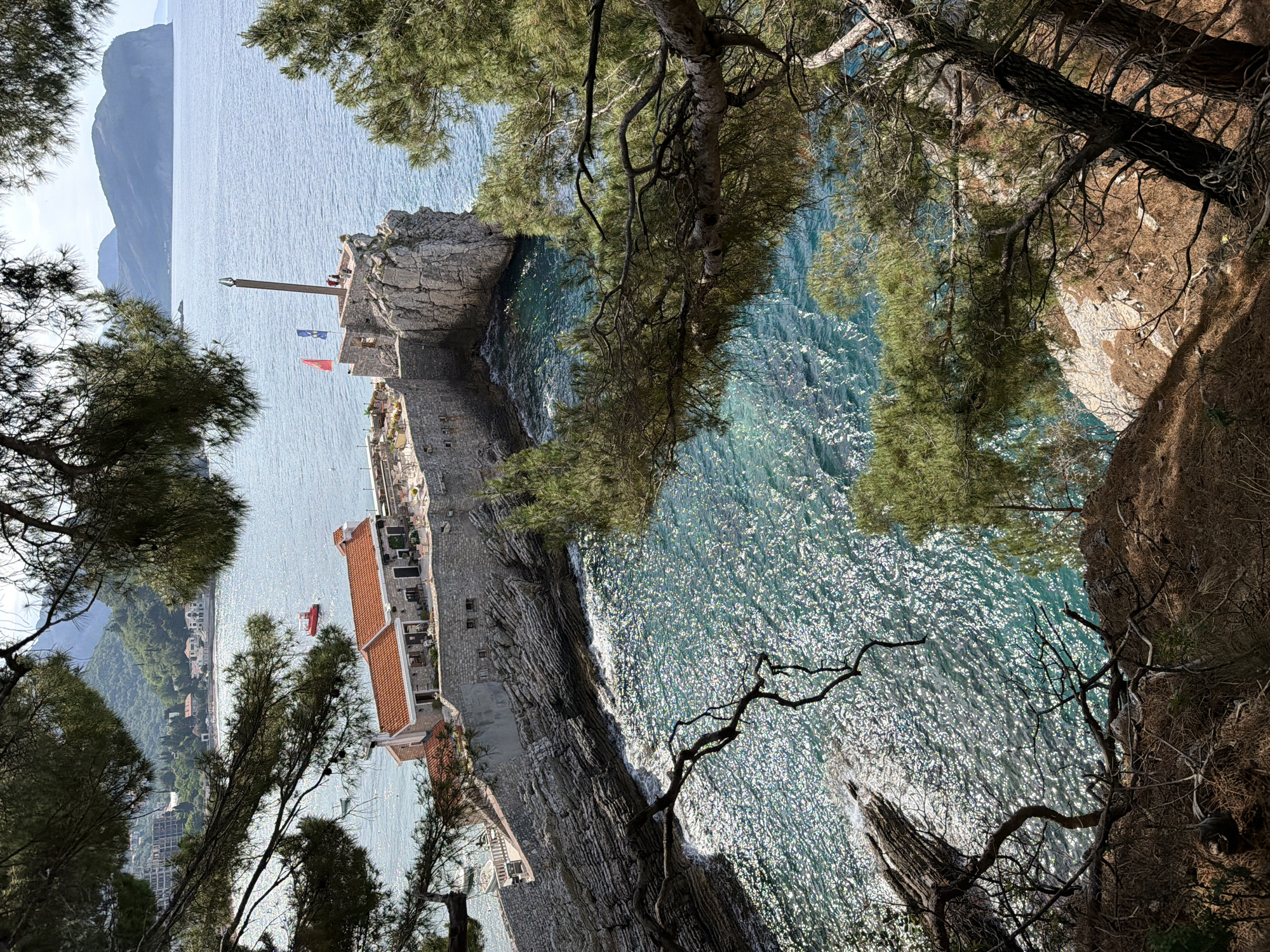 An aerial view shows Petrovac centar, Montenegro. Buildings with red roofs are situated beside the sea. An area of rock and cliff faces edge a turquoise sea, with visible tree foliage at the periphery of the image. The environment is sunlit.