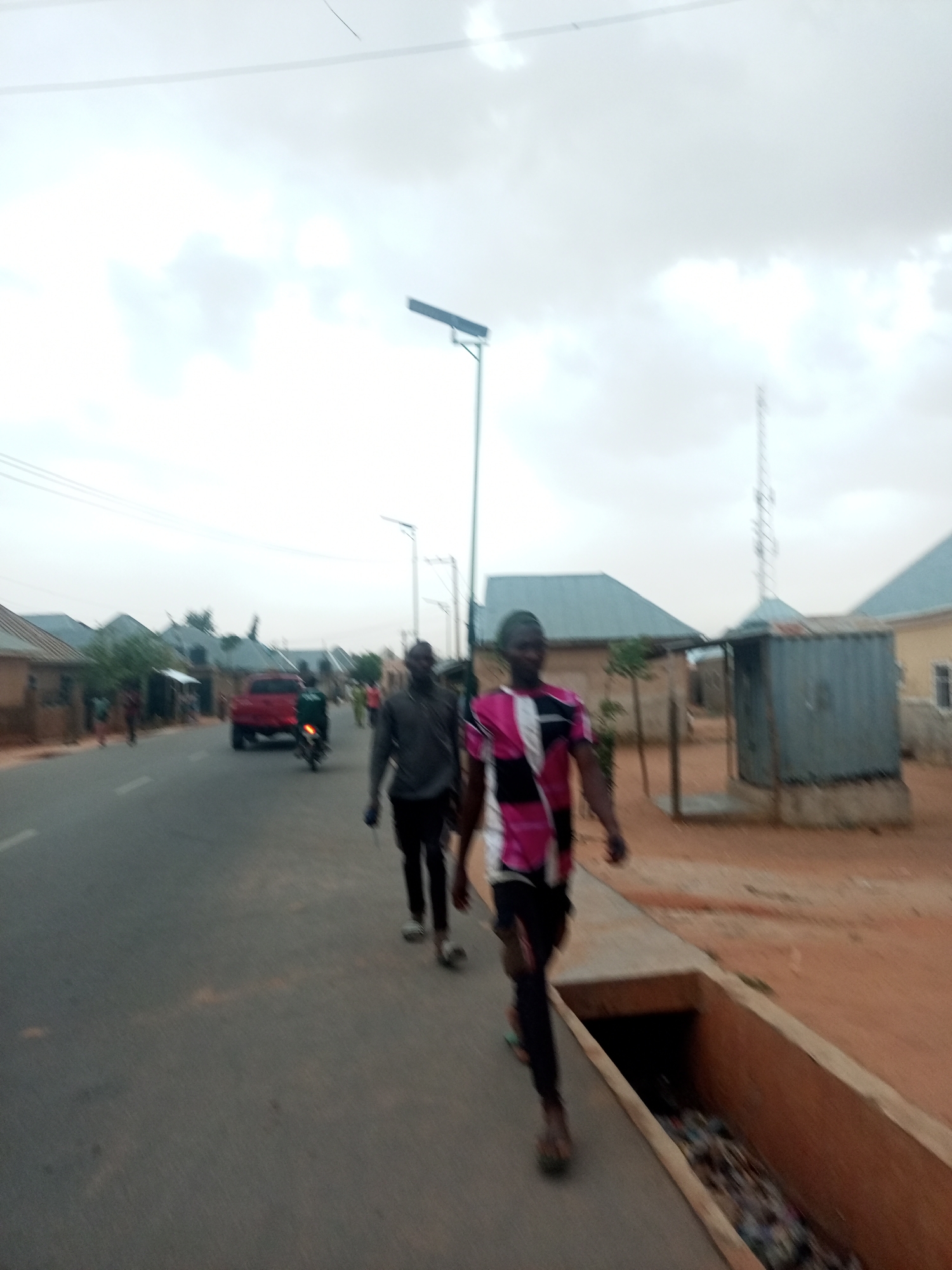 The image shows a street scene in Unknown, Unknown, featuring two men walking along the road. A red pickup truck and a motorcycle are visible in the background. A cellular tower is present in the distance. Streetlights line the road. A drainage ditch runs alongside the road, containing debris. Houses are visible in the background. The sky is overcast.