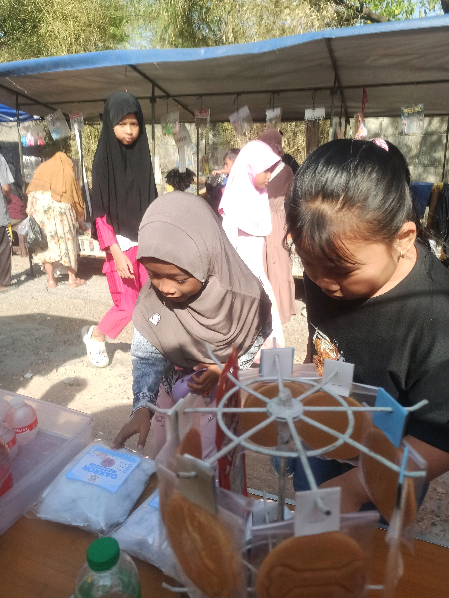 In Telagamurni, Indonesia, a market scene unfolds with four individuals, primarily young girls, near a stall. In the foreground, a girl wearing a gray hijab and a denim jacket reaches for packaged cotton. To her right, another girl in a black t-shirt looks down at a decorative Ferris wheel display of cookies. Behind them, two other girls are visible: one in a black hijab and bright pink pants, and another in a light pink hijab. The stall features a blue canopy, and various items, including packaged goods and hanging displays, are present. The ground appears to be dirt and gravel, and the background is filled with foliage, suggesting an outdoor setting.