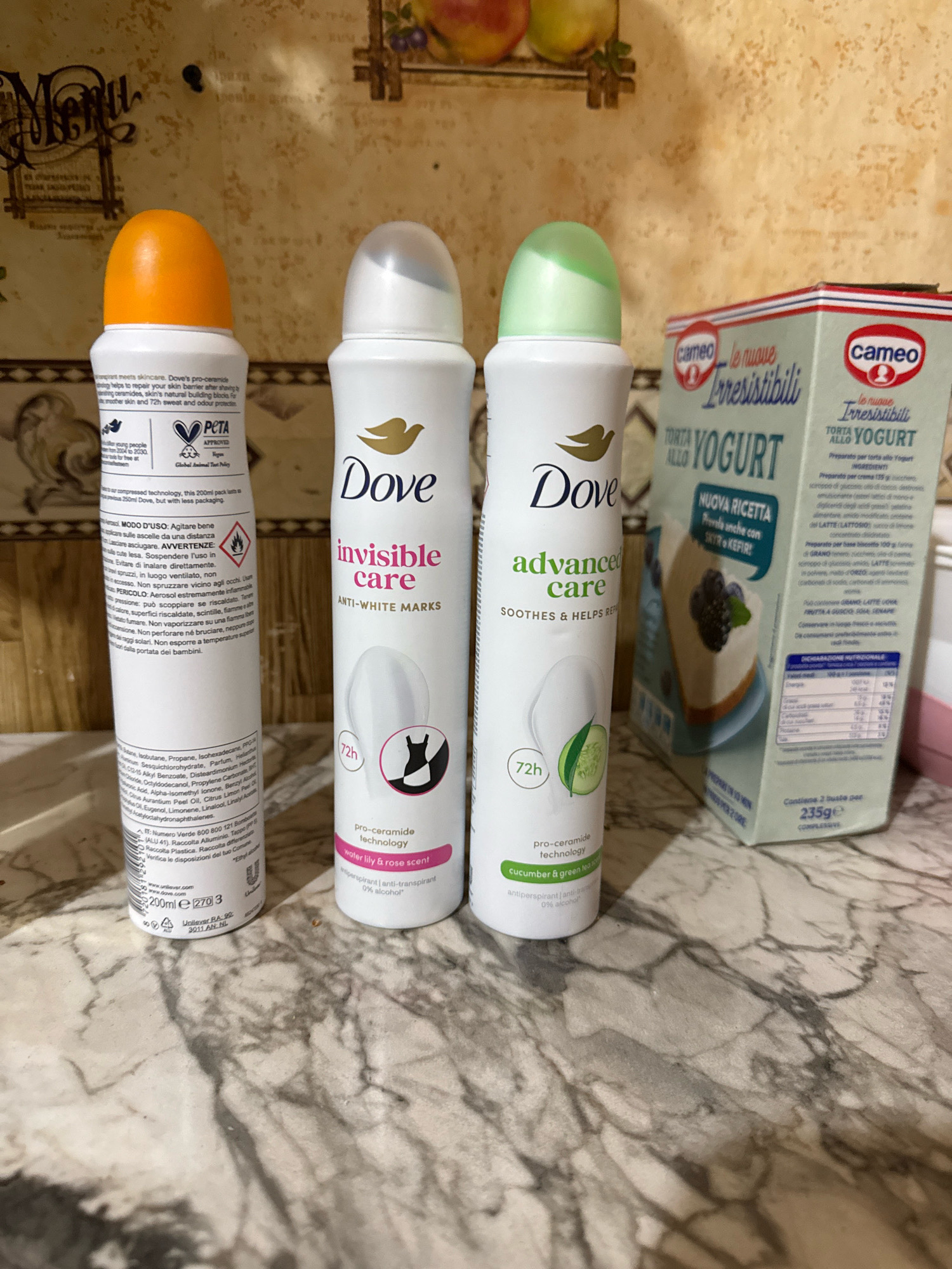 An overhead medium shot displays a marble countertop with three personal care products and a box of dessert mix. On the left is a white aerosol deodorant spray with an orange cap. To its right are two more aerosol deodorant sprays, one white with a silver cap and the other white with a green cap. Next to these sprays is a light blue box of dessert mix with a picture of a cheesecake on it. The background features a wall with a decorative pattern and a framed picture. There are no people in the image, and no actions are taking place.