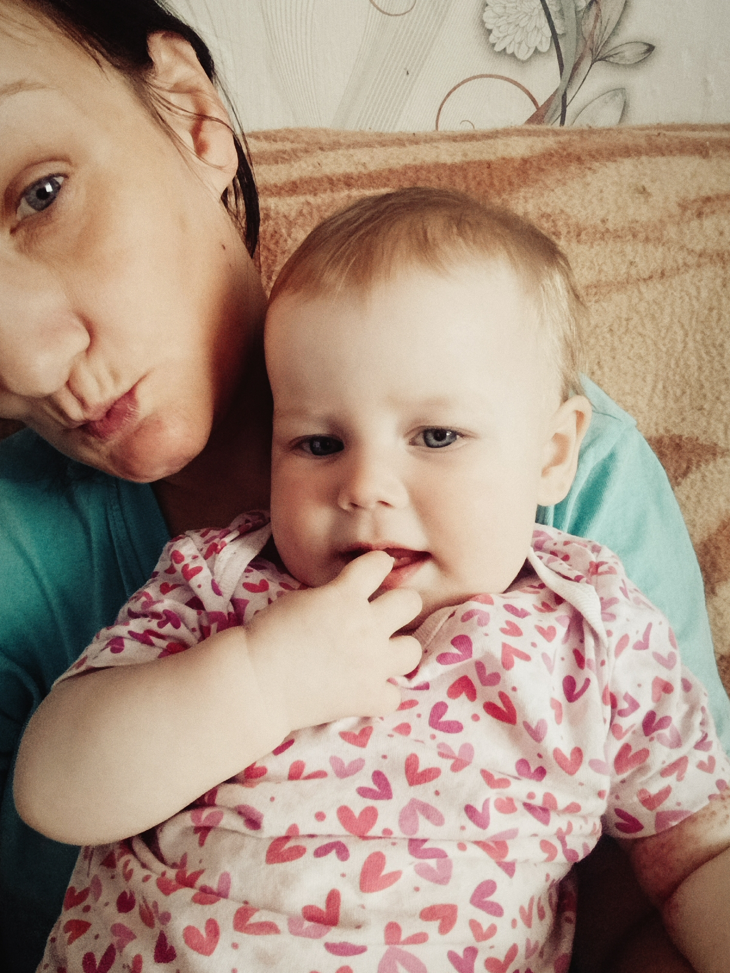 A woman and a baby are captured in a close-up shot, seemingly indoors. The woman, with light skin and dark hair, is wearing a blue shirt and appears to be making a pursed-lip expression. The baby, fair-skinned with light brown hair, wears a white onesie patterned with red and pink hearts, has their fingers in their mouth, and is being held by the woman. The background includes a brown, textured surface and a wall covering featuring a floral pattern. There is no indication of the city of Фоки, Russia, being relevant to this image.