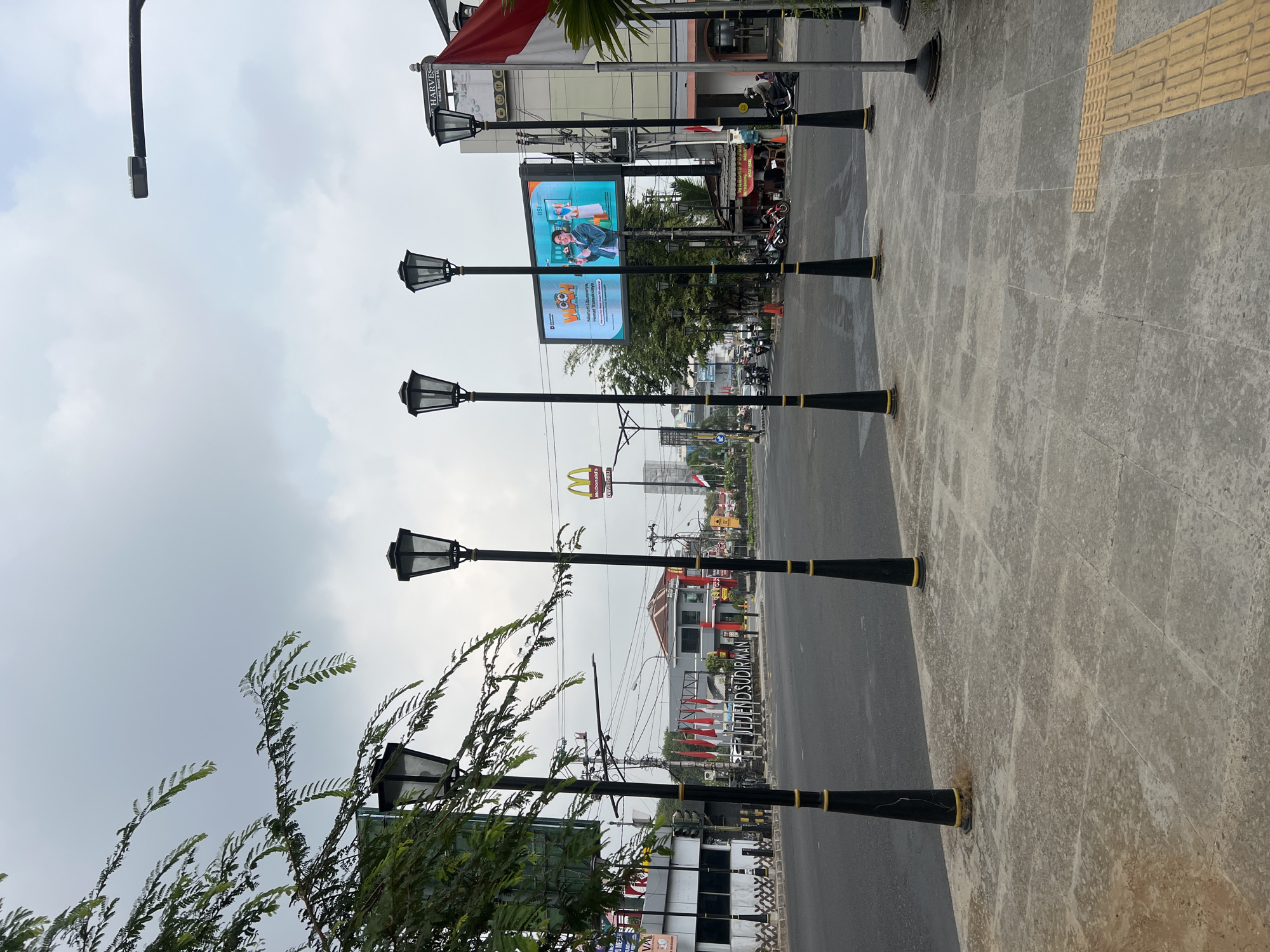 The image depicts a street scene in Yogyakarta, Indonesia, with a focus on the intersection of Jalan Jenderal Sudirman and Jalan Mangkubumi. In the foreground, a paved sidewalk is lined with decorative lampposts. Beyond the sidewalk, a wide asphalt road carries light traffic, including motorcycles and cars.
Several prominent establishments are visible on either side of the road. A McDonald's restaurant with its iconic golden arches stands out, as does a building labeled "HARVES." A large digital billboard displays an advertisement for "YOOCH" and "BSI" (Bank Syariah Indonesia). Red and white Indonesian flags are planted along the street, signifying national pride.
The sky is overcast, suggesting a cloudy day. The overall atmosphere is that of a typical urban environment in Indonesia, bustling with activity and commercial presence. The street signage indicates the location of Jalan Jenderal Sudirman, a major thoroughfare in Yogyakarta.