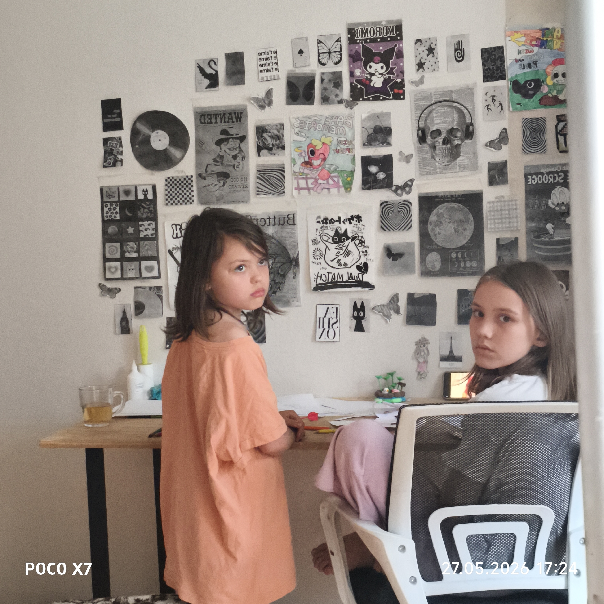 Two young girls are positioned around a desk in a room with a wall decorated with numerous paper cutouts and images. One girl, wearing an orange t-shirt, is standing, while the other is sitting in a white mesh office chair, looking directly at the camera. On the desk, there are papers, writing utensils, a glass of liquid, and a small decorative item. The wall behind the girls features a collage of black and white images, including depictions of butterflies, skulls, and various abstract shapes. A smartphone is placed on the desk behind the sitting girl. In the lower left corner of the image, the text "POCO X7" is visible, and in the lower right corner, the timestamp "27.05.2026 17:24" is displayed. The location is Unknown, Unknown.