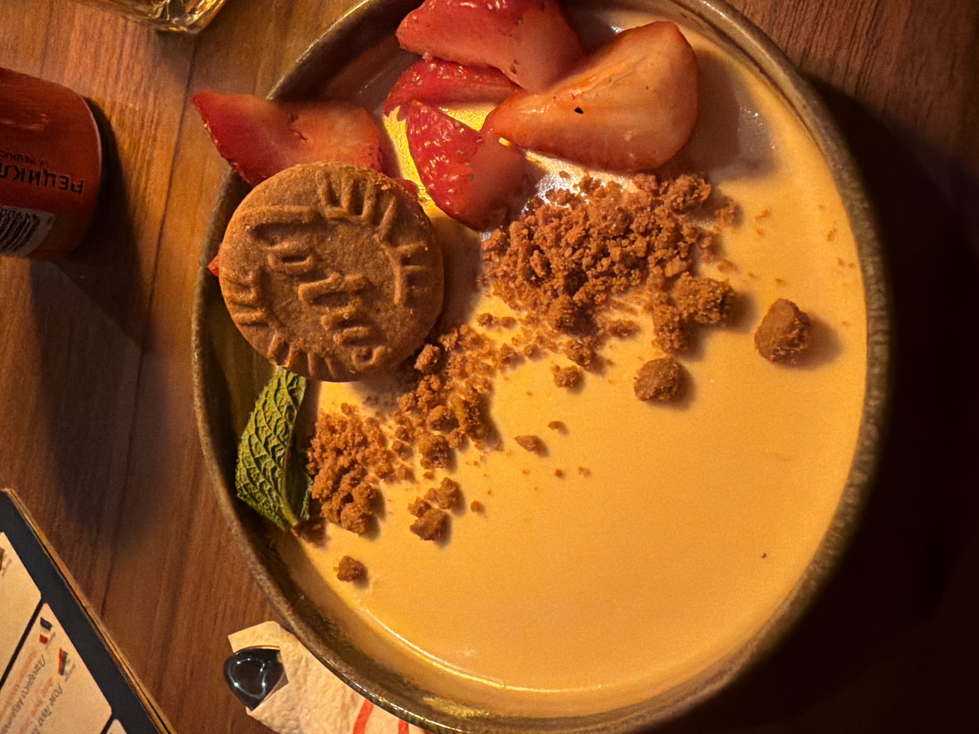 The image captures a dessert dish presented on a dark wooden surface. The dessert features a golden-hued, creamy base, likely a custard or pudding. Adorning the creamy surface are sliced strawberries, a round biscuit with a decorative pattern, and a scattering of crumbled topping. A mint sprig is positioned near the biscuit. The edge of a menu or card is visible in the lower left, with some text in a foreign language. A small container or bottle and a wrapped object are partially visible in the upper left and lower center of the frame, respectively. The setting suggests an indoor dining environment, likely in a restaurant or cafe. The location is specified as Sofia, Bulgaria.