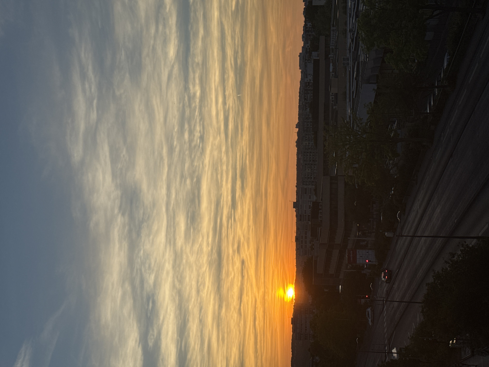 This is a vertically oriented, undated, and uncaptioned photograph that captures a city scene at sunset. The left side of the image is dominated by the sky, which transitions from a pale blue at the top to a vibrant orange as it nears the horizon. Wispy, elongated clouds streak across the sky, catching the warm hues of the setting sun. A small, white object, possibly an airplane, is visible in the sky.
On the right side of the frame, a cityscape rises against the colorful sky. Buildings of varying heights and architectural styles are visible, some appearing to be residential or office blocks. Trees with dark green foliage line the street, creating a contrast with the urban structures. A road runs through the scene, with streetlights and traffic signals visible. A few cars are on the road, one of which has its brake lights illuminated. The scene is dimly lit, indicating either the early dawn or late dusk. The overall mood is serene and peaceful, characterized by the beautiful sunset and the quietude of the urban environment. There is no discernible text in the image.